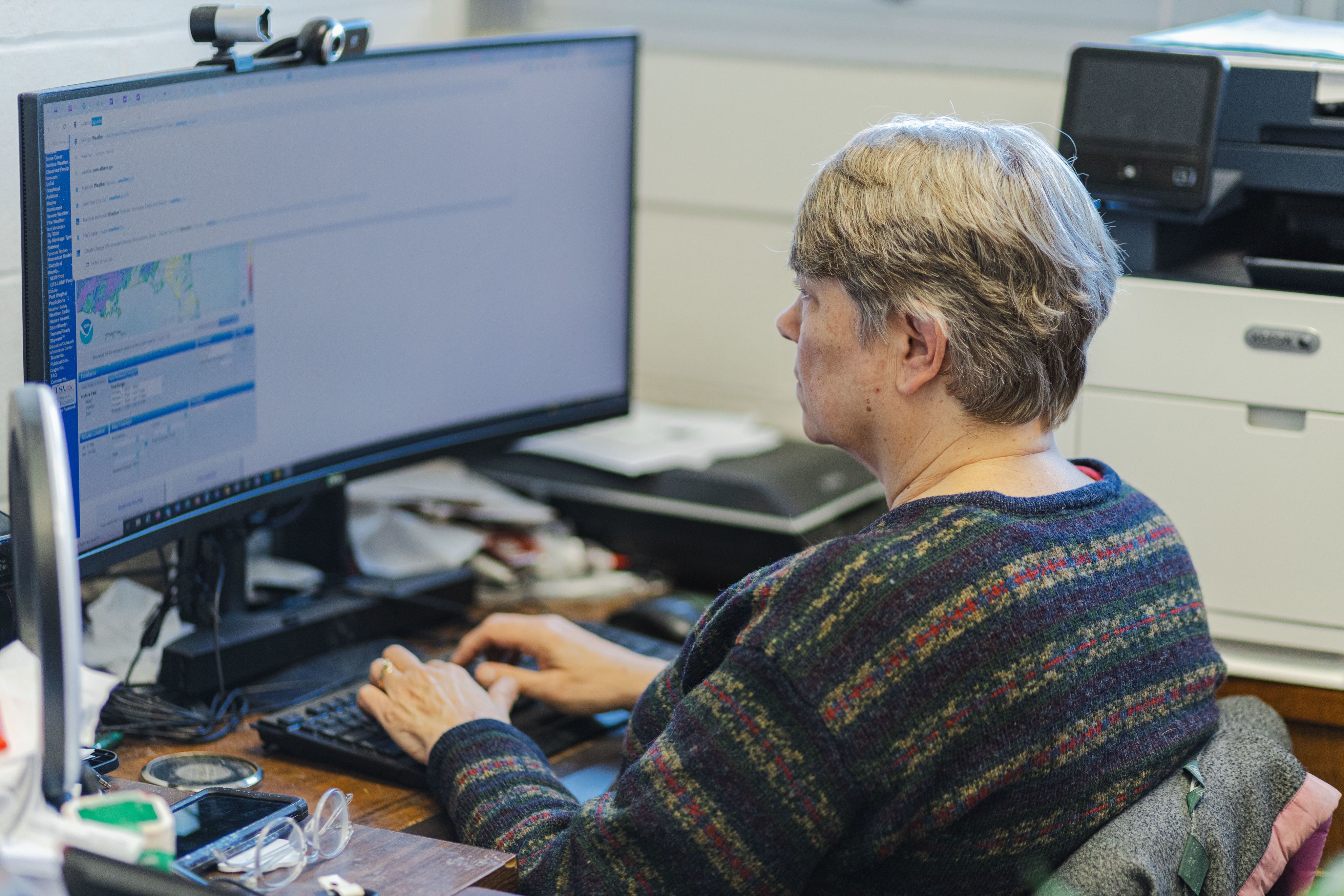A woman with short hair, wearing a blue striped sweater views weather data on a large, curved computer screen
