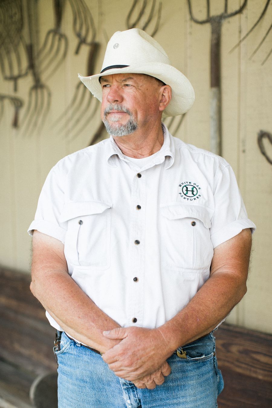 Will Harris stands with his hands folded in front of him wearing a white cowboy hat, a white button up shirt withthe White Oak Pastures logo on the chest, and blue jeans.