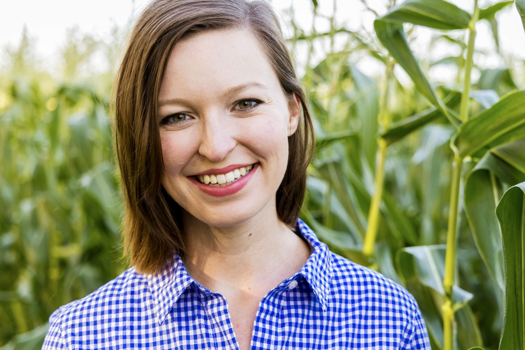 A woman with shoulder length brown har and a blue and white gingham checked shirt smiles while standing in a corn field.
