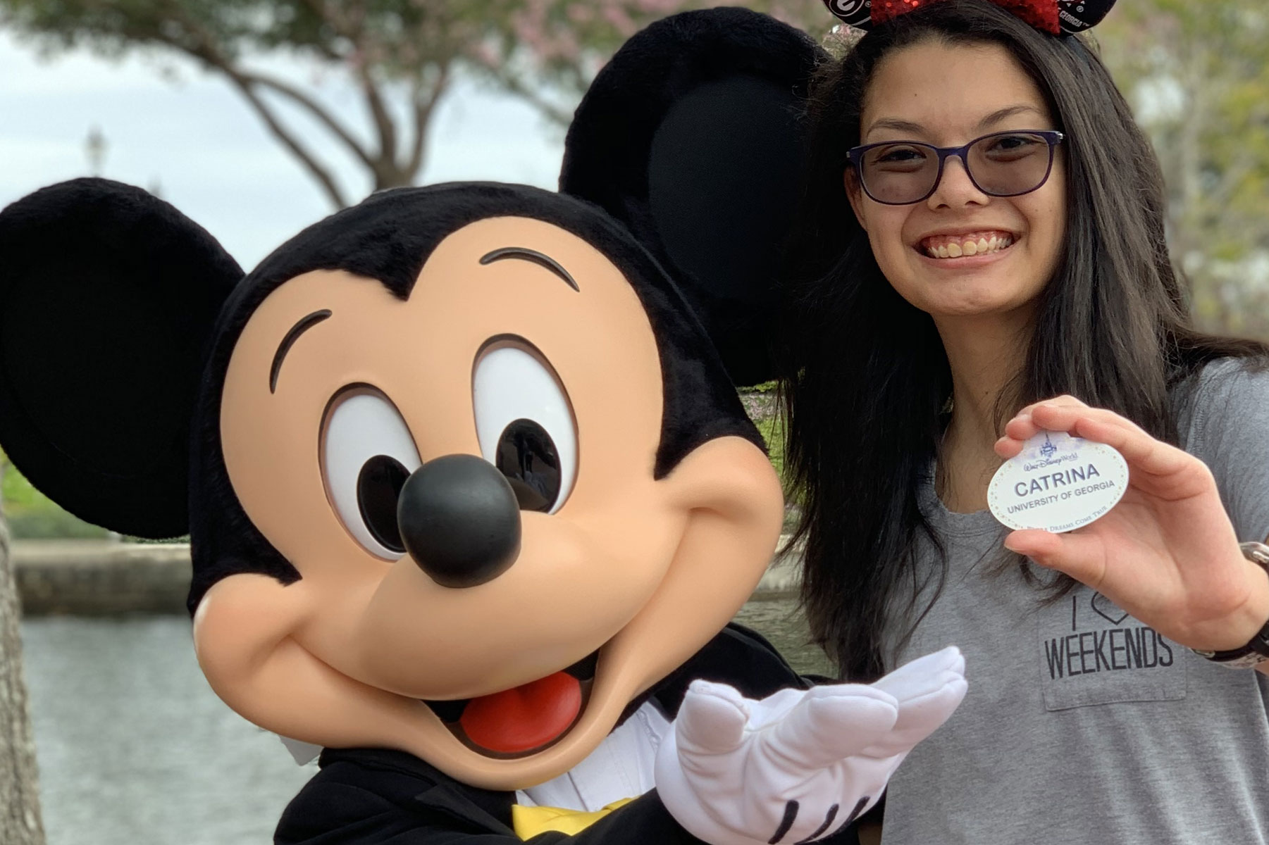 A smiling student holding up a nametag poses with a Mickey Mouse character