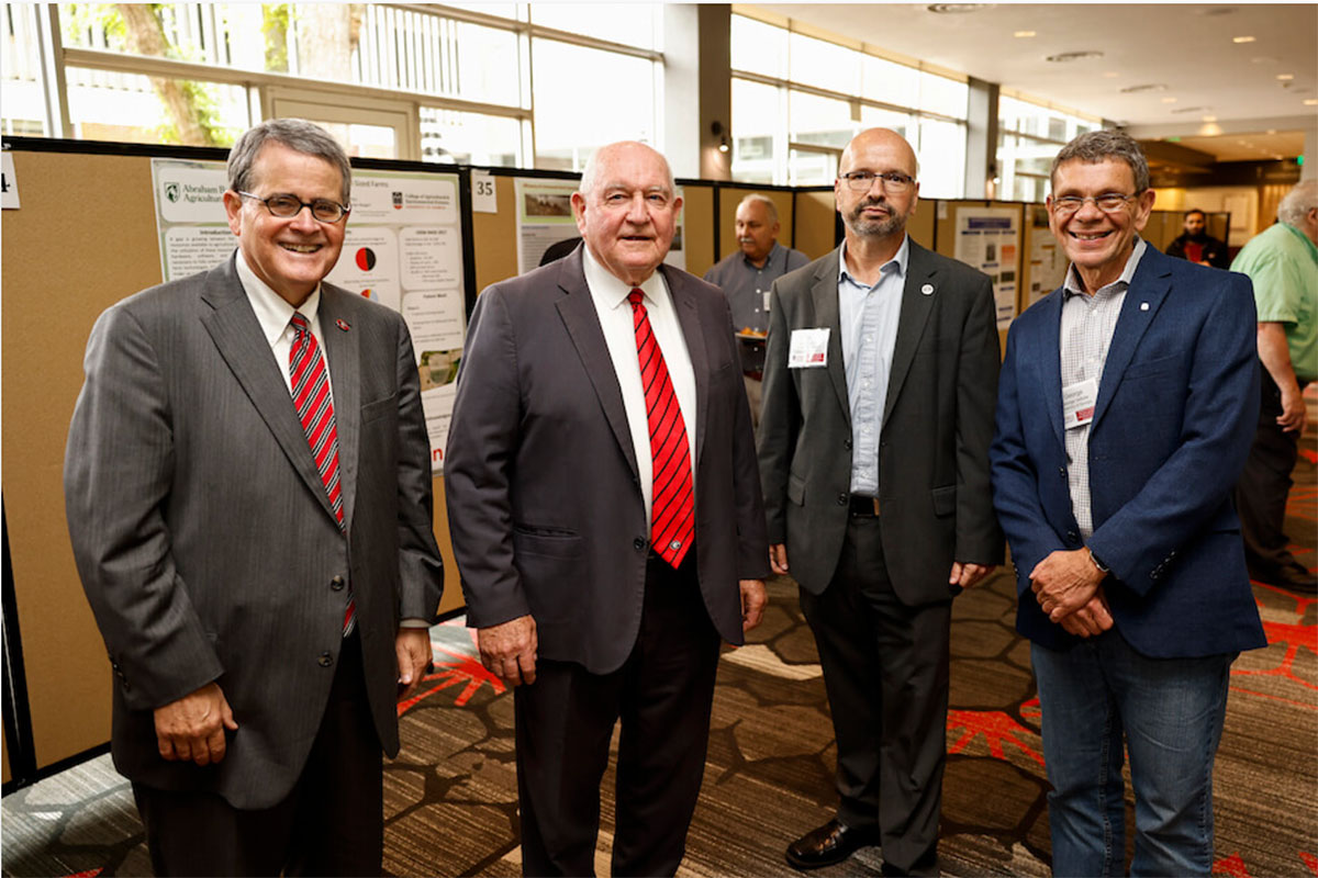Four men in business attire attend a conference with research posters hanging behind them, smiling together