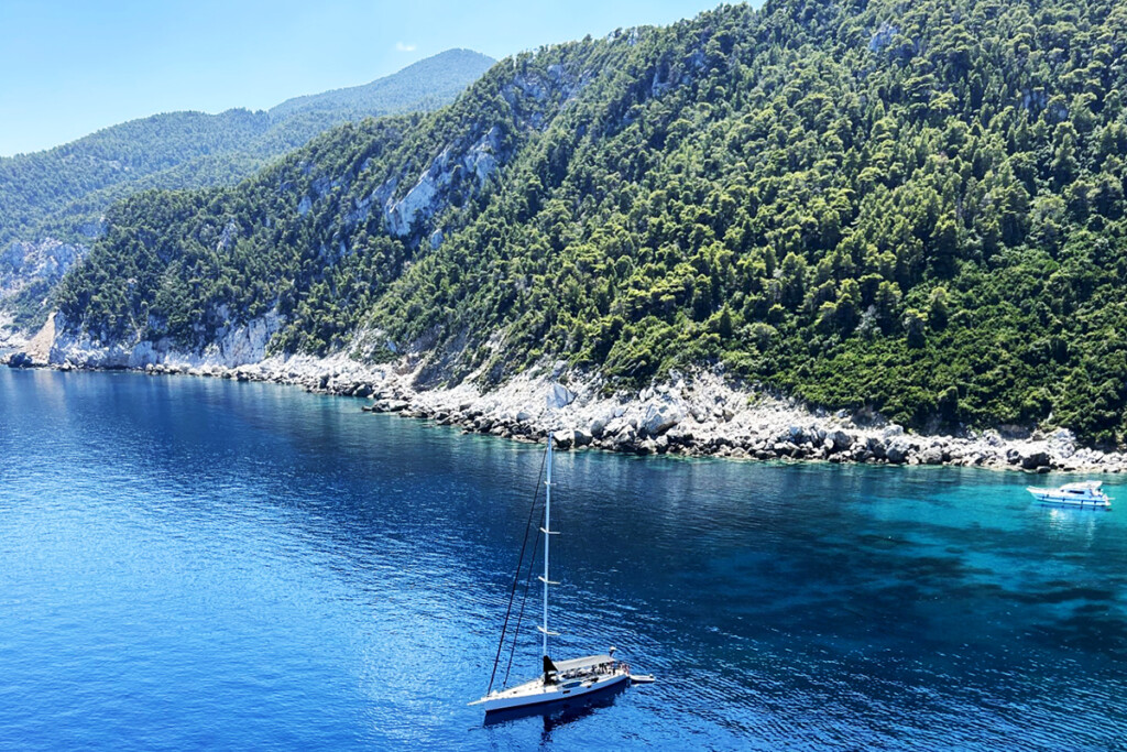 Sailboats on clear blue water along a rocky, forested coastline in Greece.