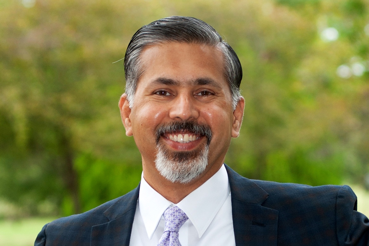 A man with salt and pepper hair and a beard wearing a suit jacket and tie smiles at the camera.