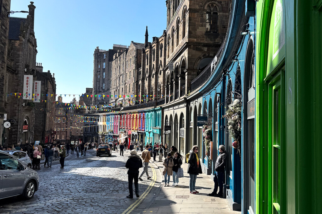 Tourists explore a street featuring colorful shop fronts under a blue sky in Scotland. 