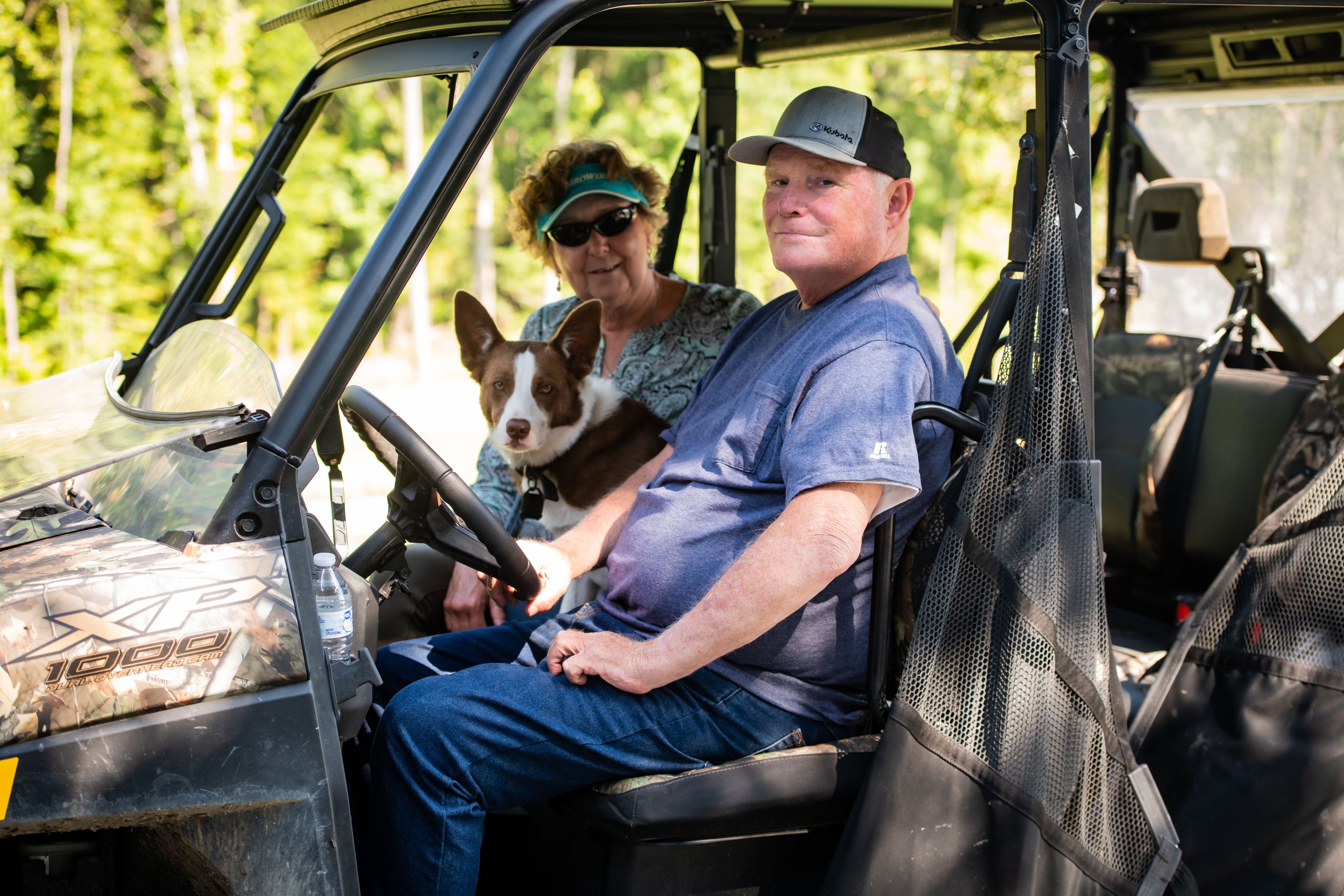 A man and a woman sit in an ATV with a brown and white cattle dog between them.