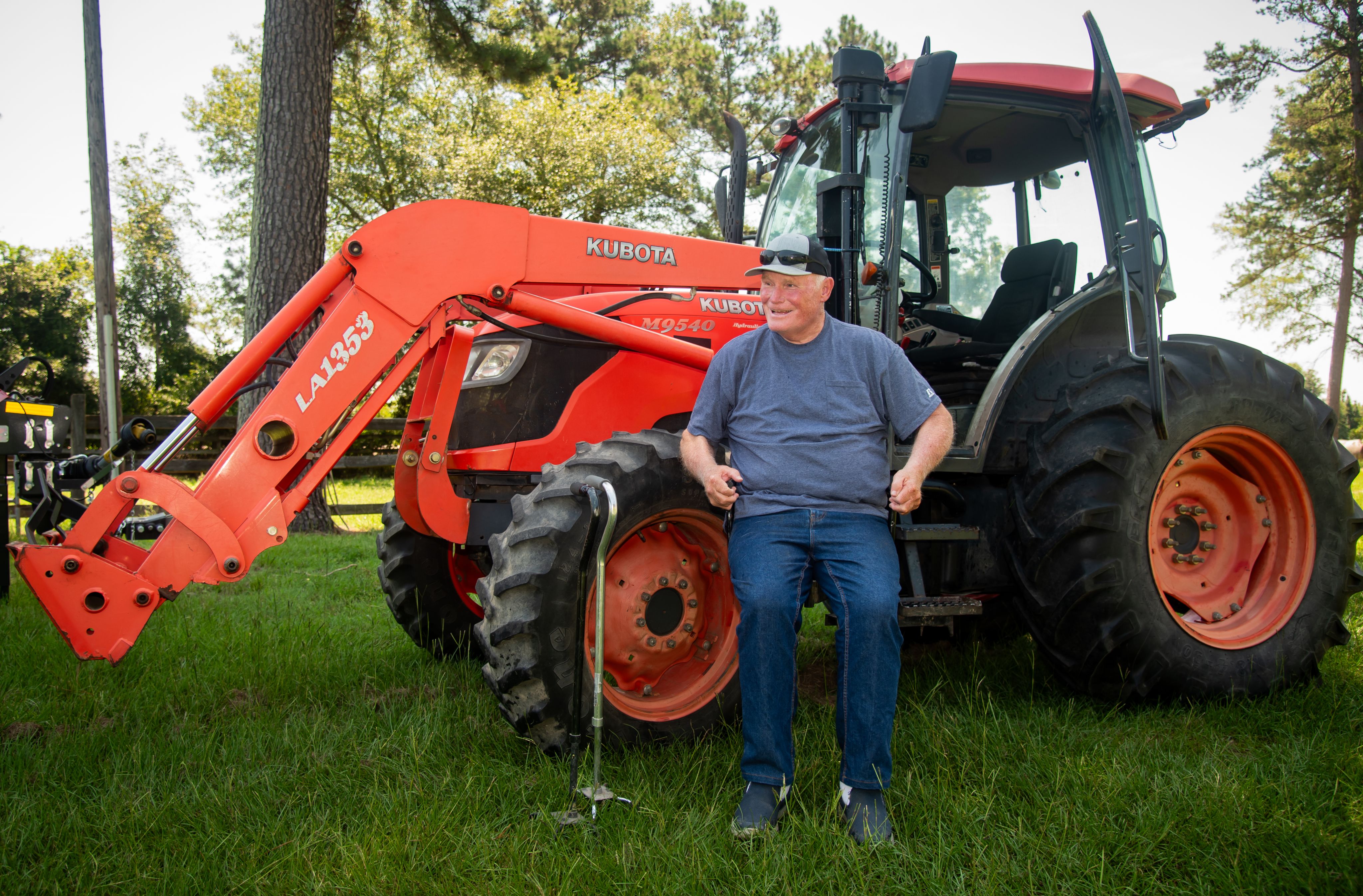 A man wearing denim jeans and a blue T-shirt sits on a lift in front of an orange tractor.