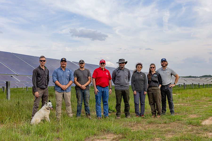 Eight people standing in front of a field of solar panels under a partly cloudy sky, with a white dog sitting on the grass to the left; the group is dressed in casual outdoor clothing, including one person in a red shirt and cap.