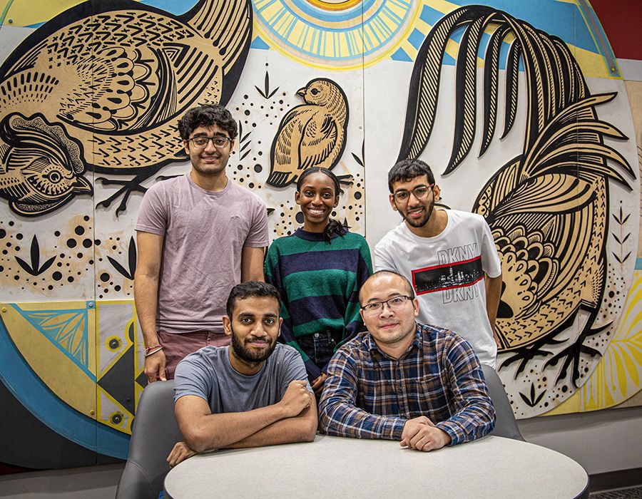 Five people pose in front of a mural featuring stylized birds, including a rooster, with intricate patterns and designs.