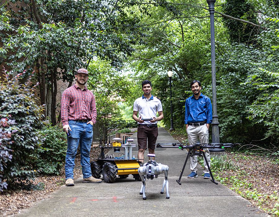 Three people standing on a wooded path with a wheeled robot, a quadruped robot, and a drone nearby