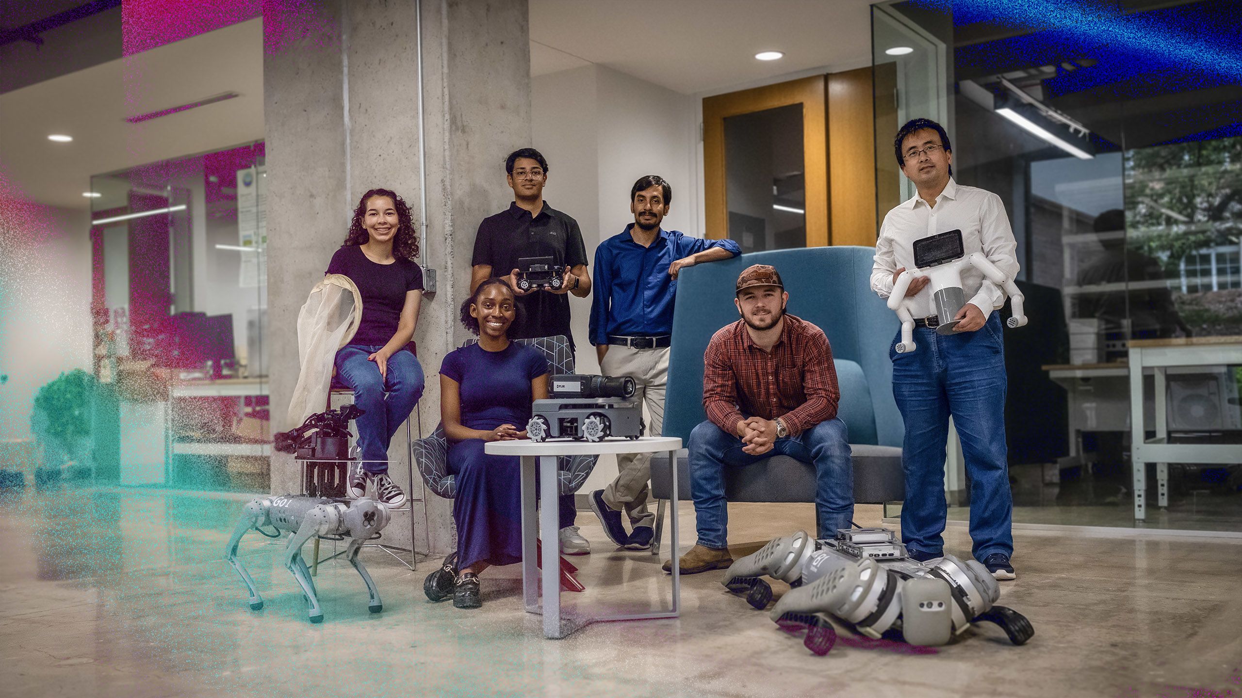 Six people in an indoor space posing with various robotic devices. One person sits on a stool holding a net next to a robotic dog. Another sits at a table with a small robot in front. Two stand holding different robots—one wheeled, one with arms. A fifth person is seated in a large chair, while the sixth stands beside them, hand resting on the chair.