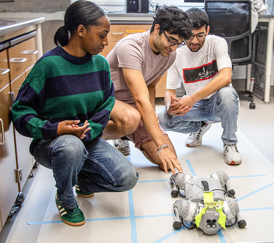 Three people kneeling on a lab floor with a robotic dog that has a gray and yellow body, surrounded by blue-taped gridlines, appearing engaged in testing or discussion about the robot’s movement.