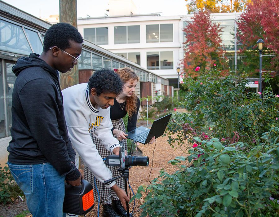 Three people in a garden setting working with equipment, a camera on a tripod, and a laptop, with greenery and a building in the background.