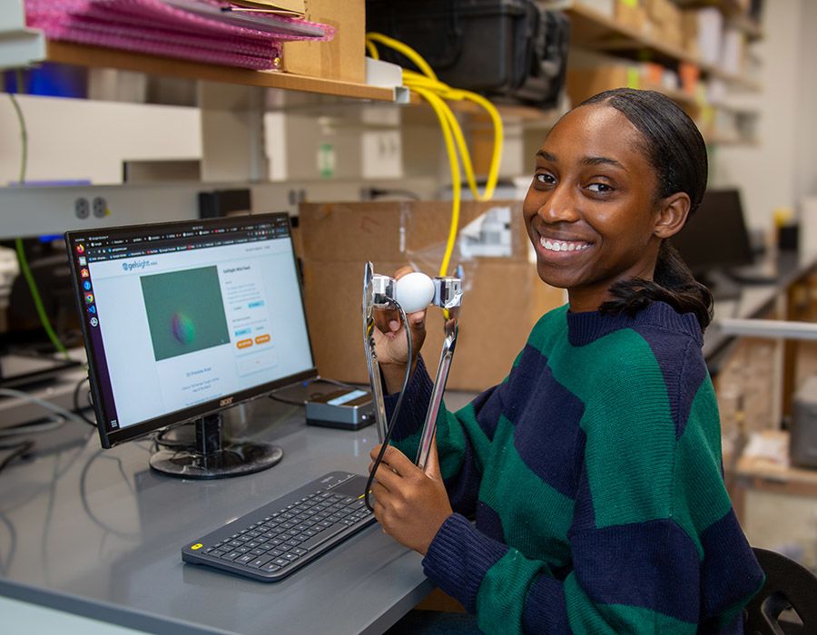 Alt text: A person seated at a desk in a lab-like workspace, holding a set of modified tongs with a white egg clasped between its two metal arms.