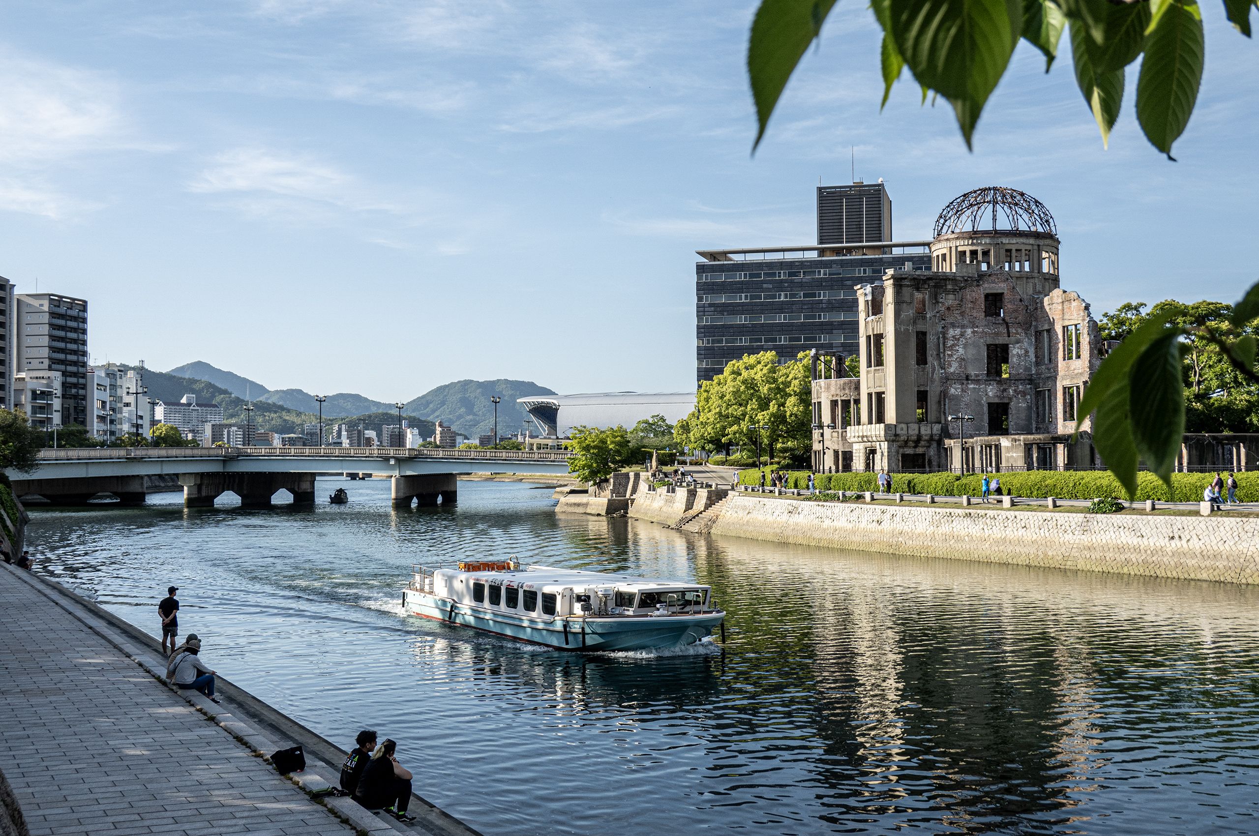 The Genbaku Dome stands by a wide river where a boat cruises, with people on the bank, and a bridge leading to distant mountains under a clear sky.