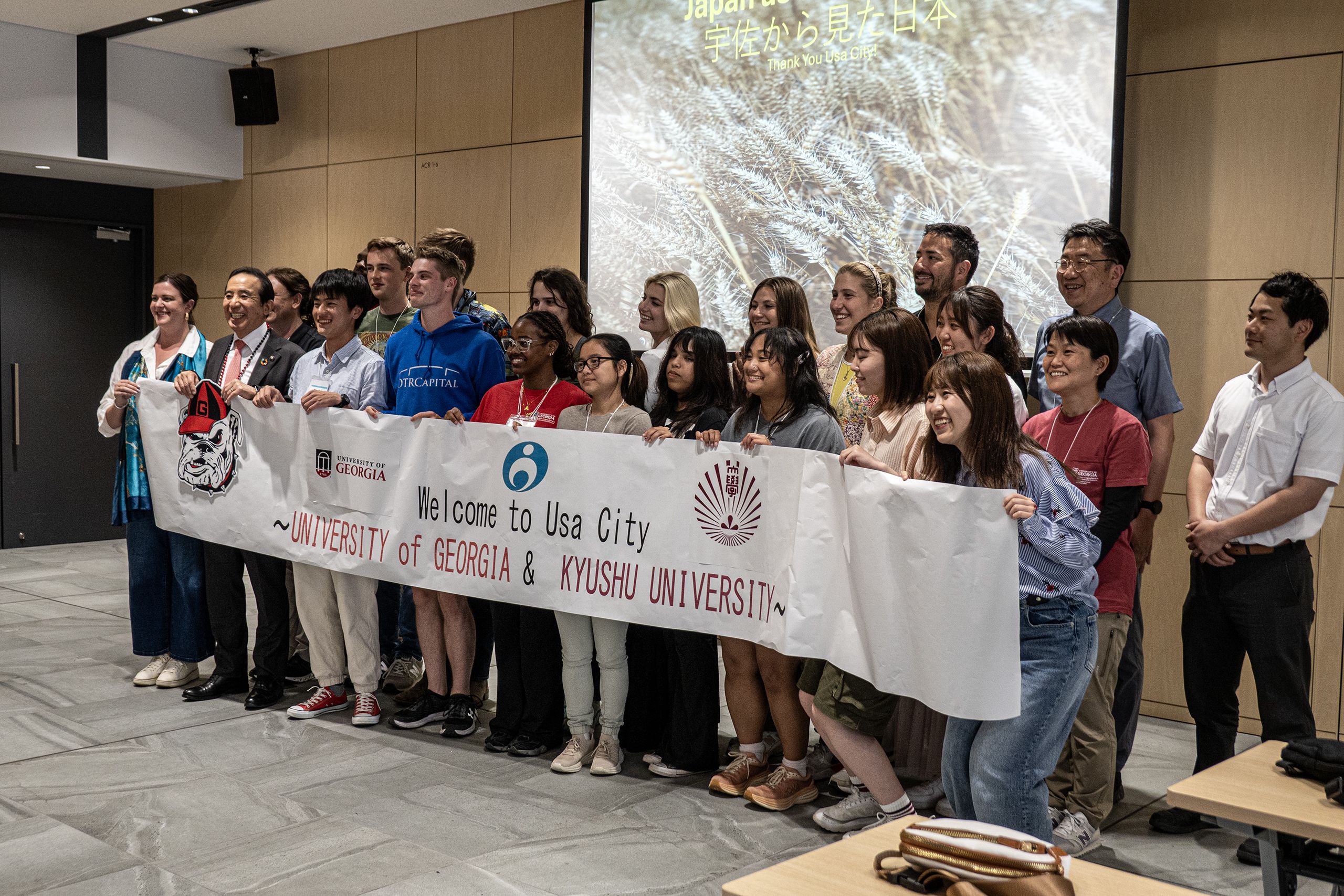 A smiling group, including students, holds a "Welcome to Usa City ~University of Georgia & Kyushu University~" banner in a room with a projector screen.