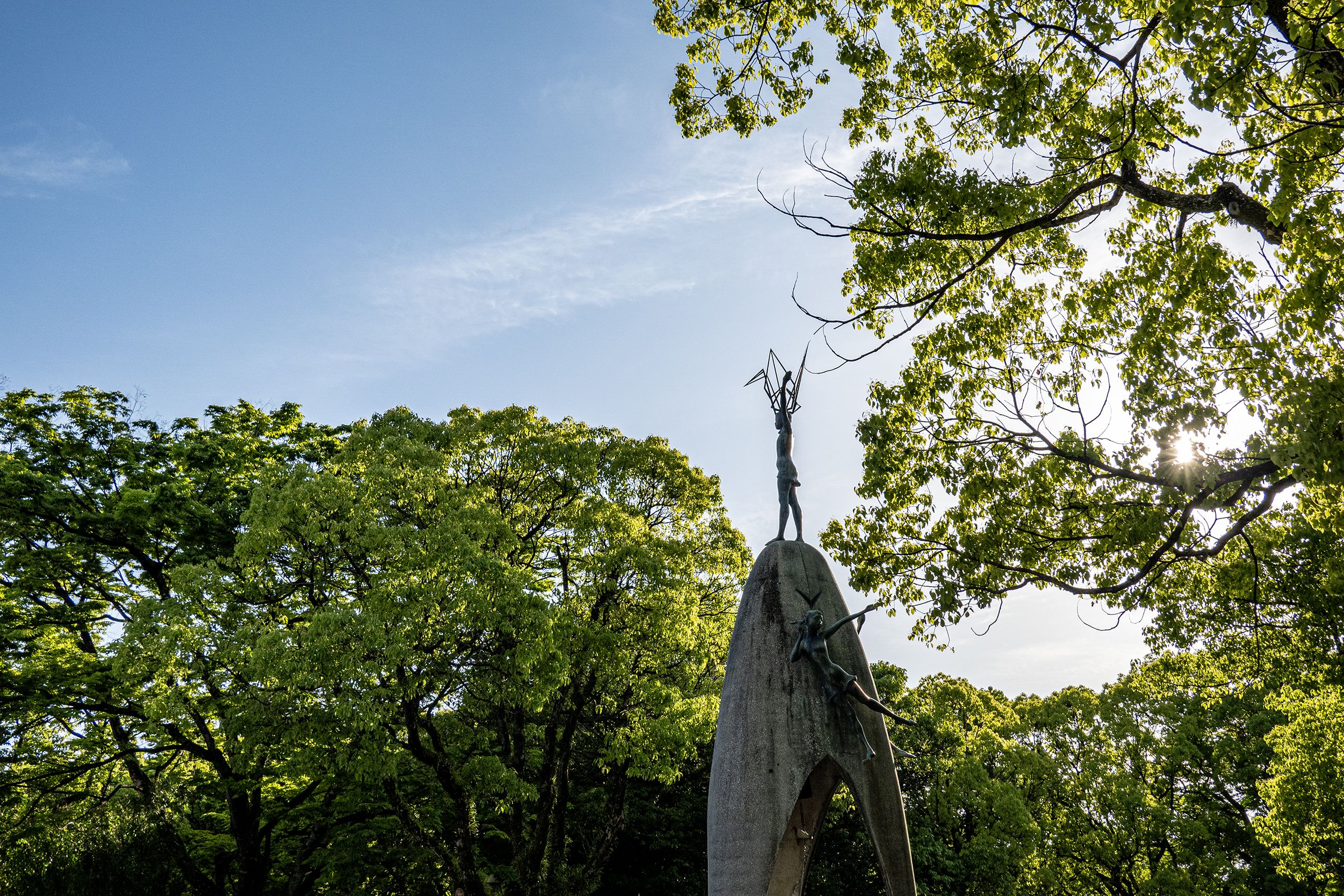 A tall stone monument with a figure reaching skyward stands among lush green trees, bathed in sunlight under a blue sky.