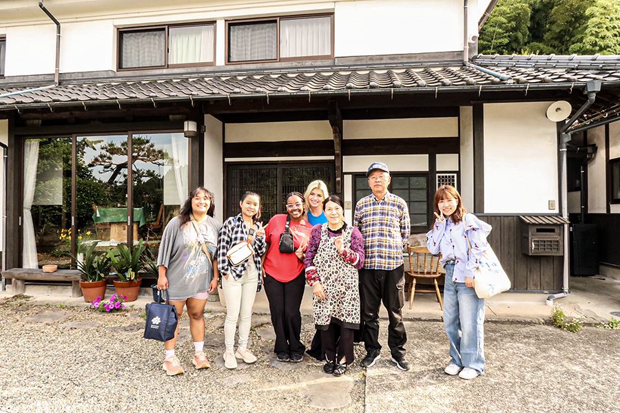 A smiling group of seven, including a host family and guests, stands in front of a traditional Japanese-style house.