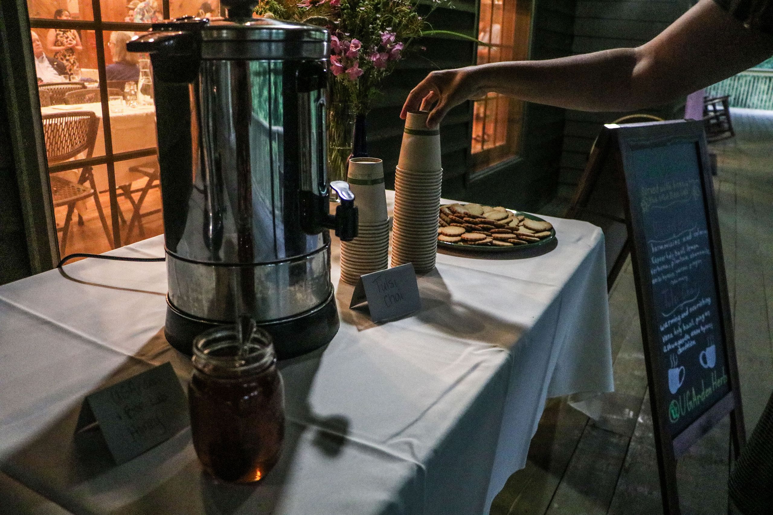 An evening tea service set outside has a metal carafe of herbal tea with a jar of honey and stacks up cups and a tray of thin wafer cookies.