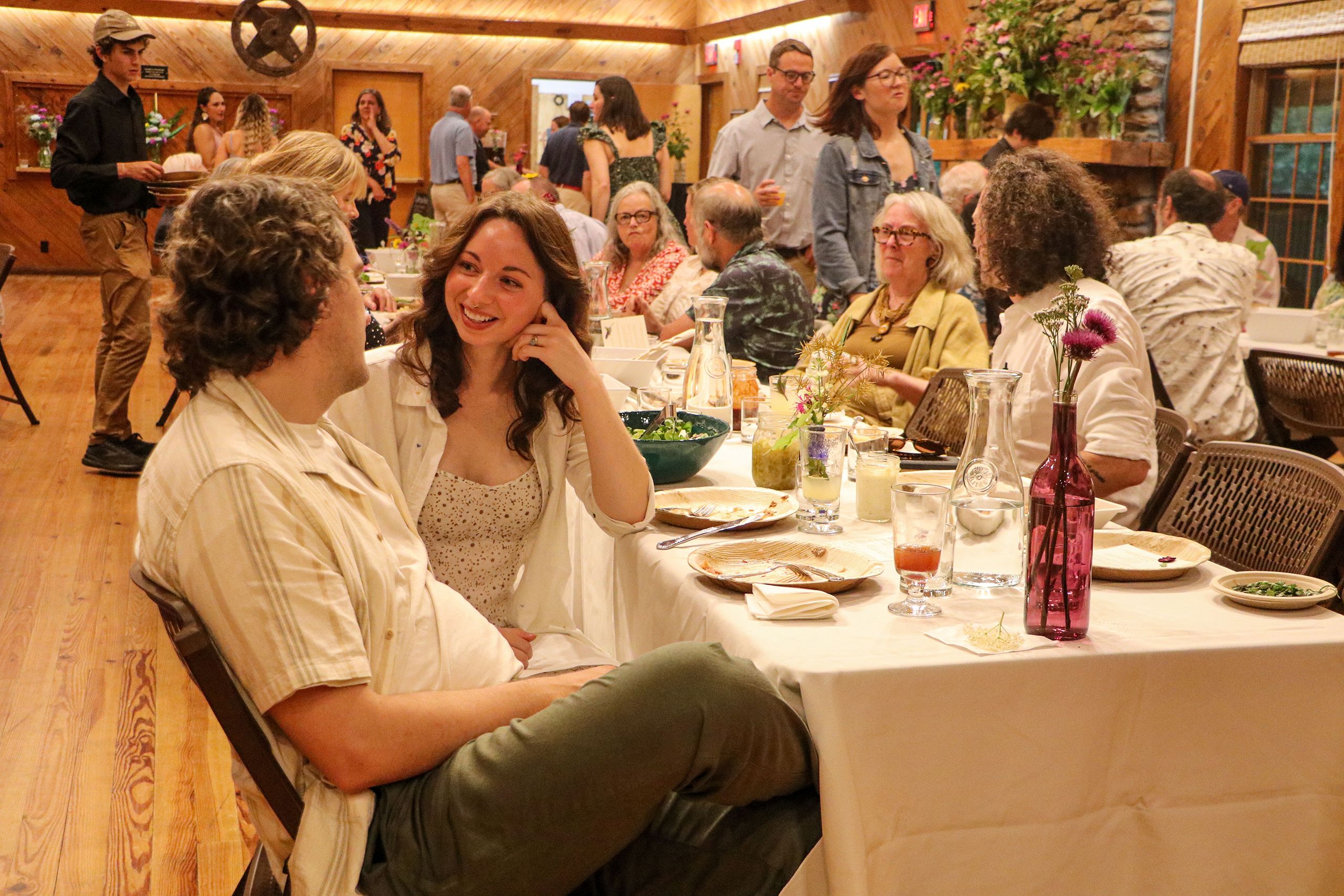 People talking as they finish dinner while sitting at long tables still covered with colorful food and drinks. 