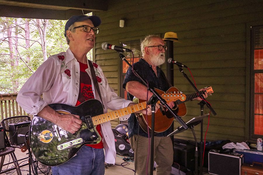 Two men with glasses and guitars are singing into microphones on a covered porch.