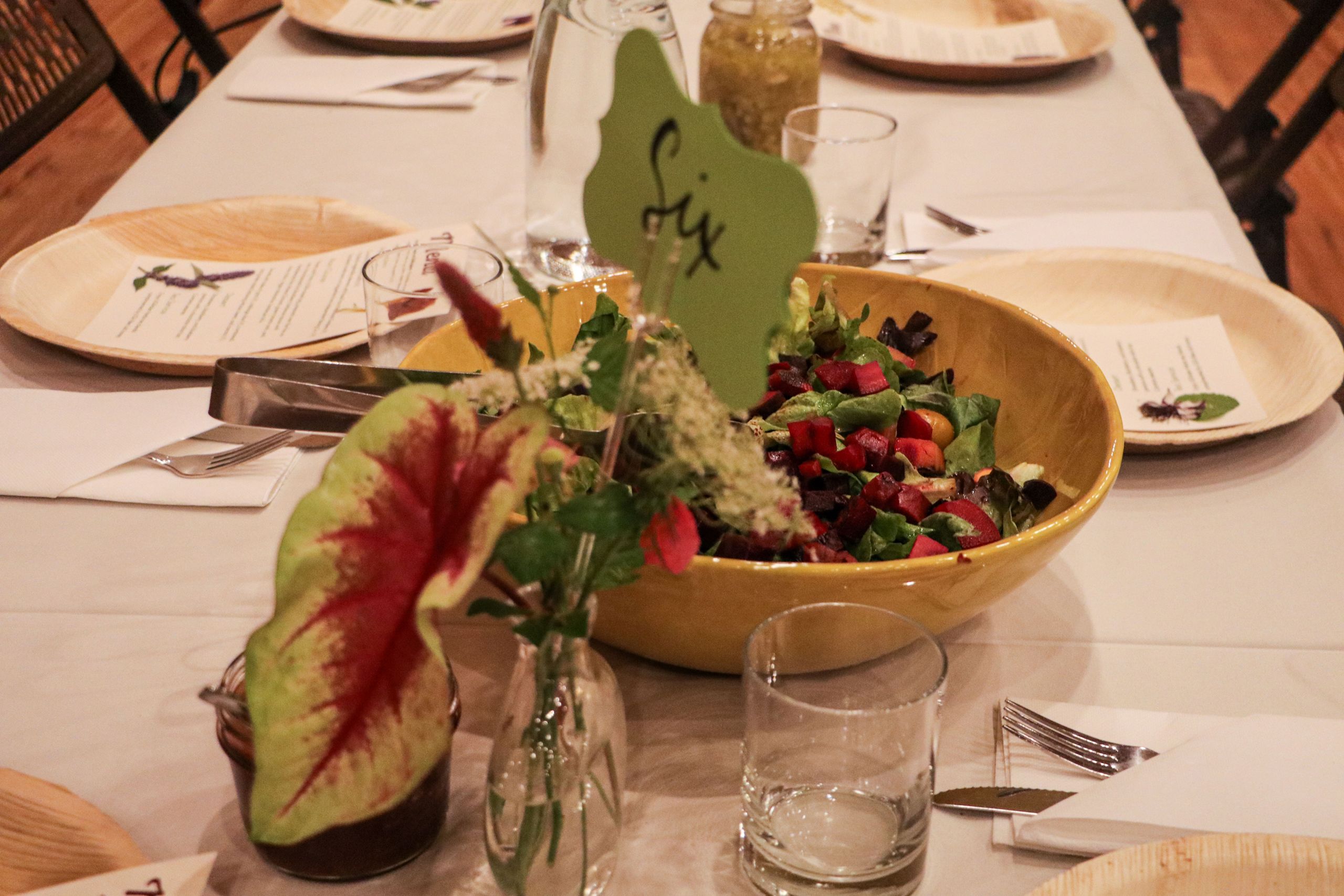 A bowl of green salad topped with red beets with flower arrangements in the foreground and place settings in the background.
