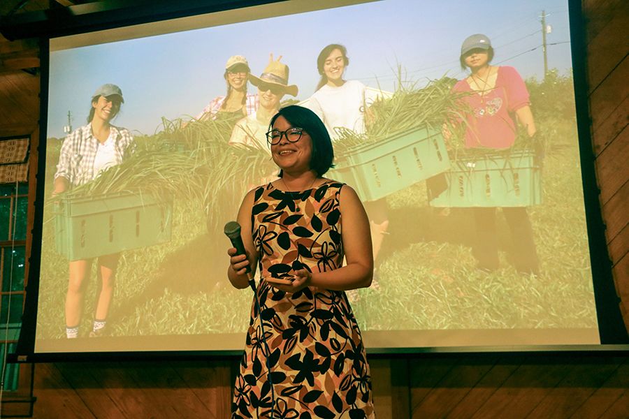 April McCoy hold a microphone and smiles as she gives a speech at the gala in front of a projector displaying photographs of the garden and volunteers over the course of 15 years. 