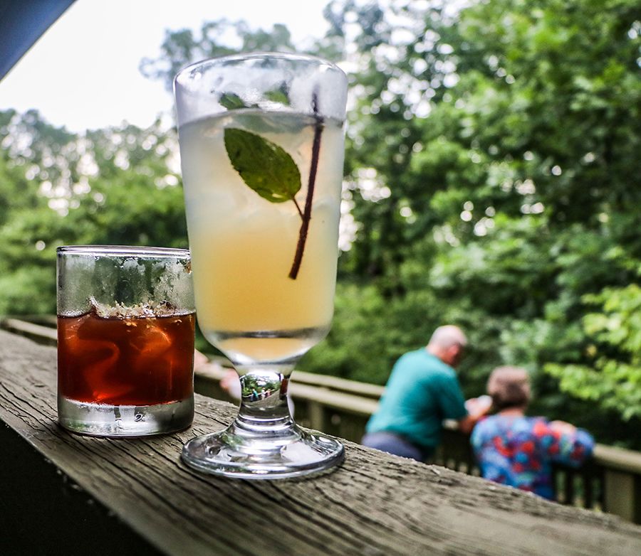 A close up image of two specialty drinks served at the banquet are resting on the porch banister. Two people can be seen in the background on the lower porch with trees behind them. 