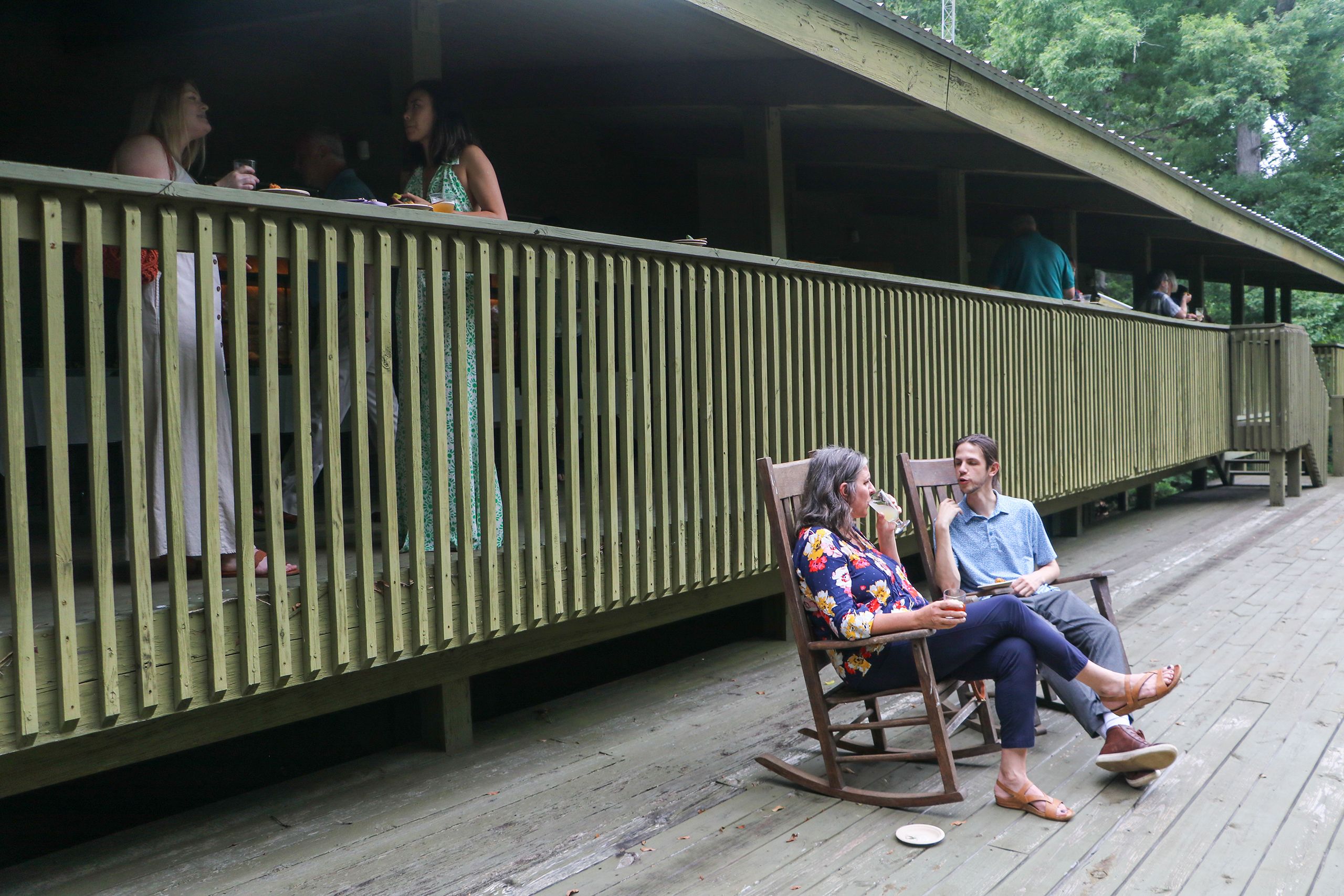 Two people sit in rocking chairs and talk while sipping drinks on a wooden deck. Another deck railing is seen in the background with vertical posts.