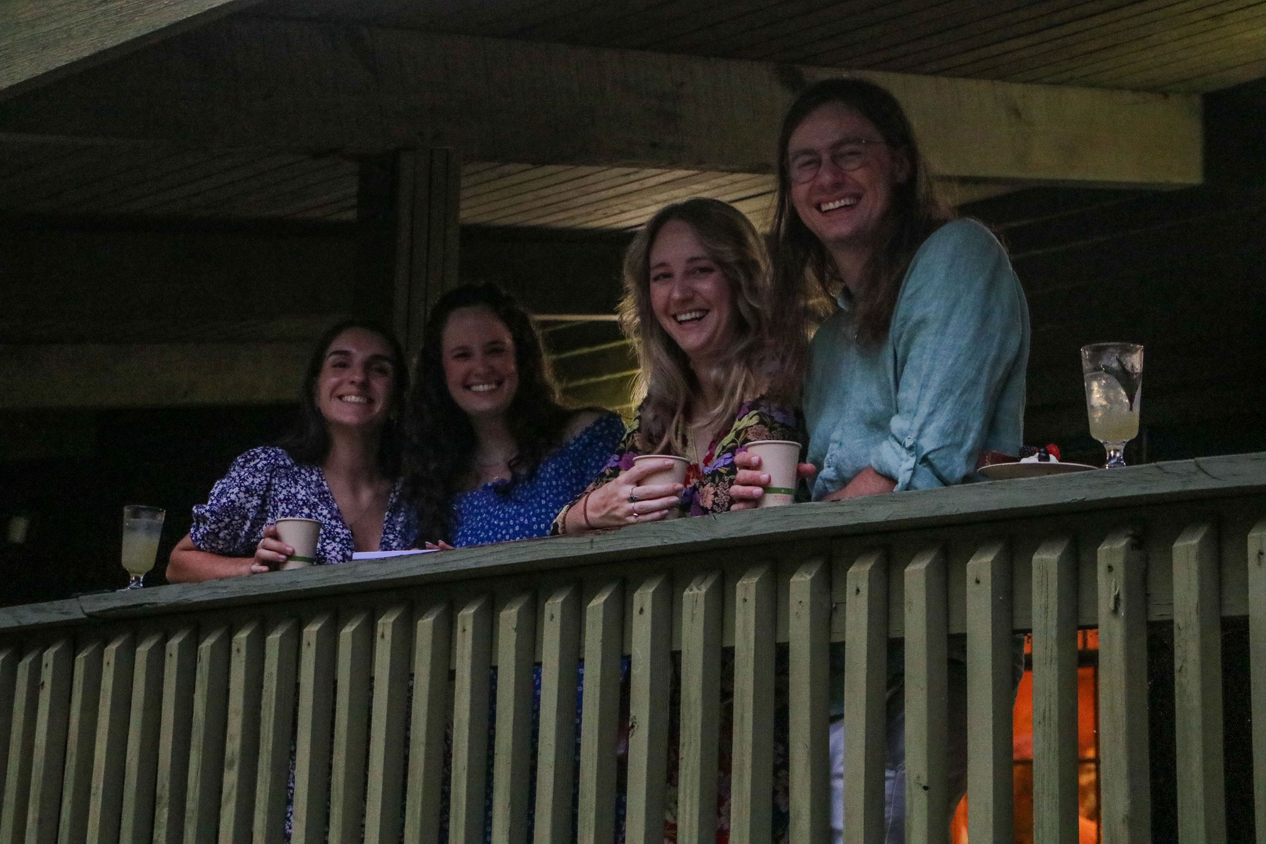 Several guests are lined up under the covered porch. They hold drinks and lean against the banister, smiling at the camera.