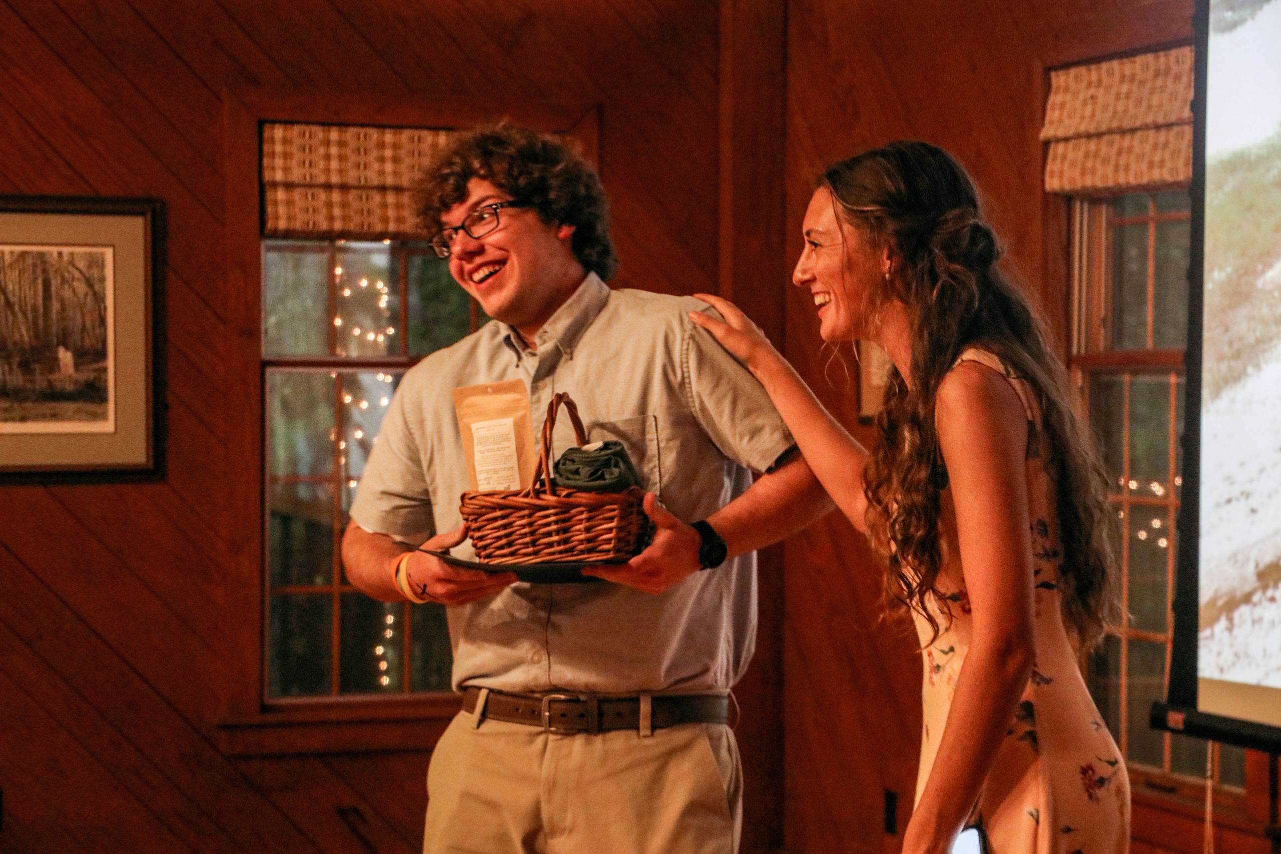 A young man smiles while holding a basket of tea and other goodies as a woman laughs and touches his shoulder. 