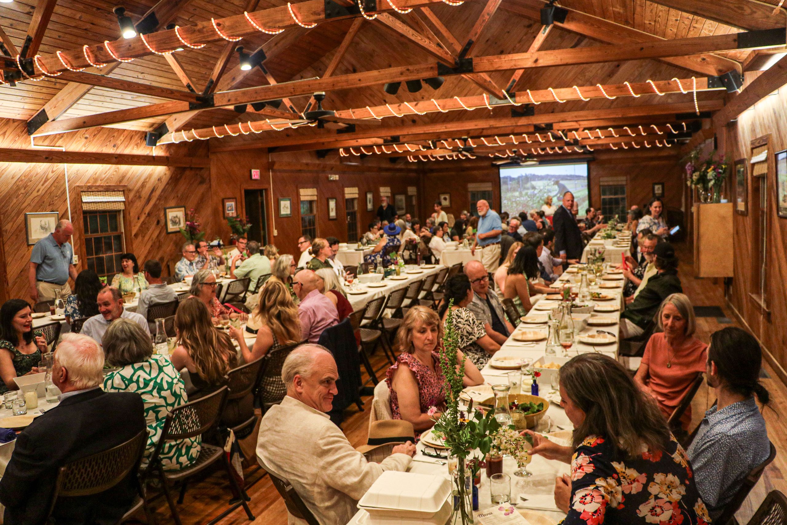 An overview shot of the banquet hall shows several long tables, with guests seated and talking as they wait for dinner. 