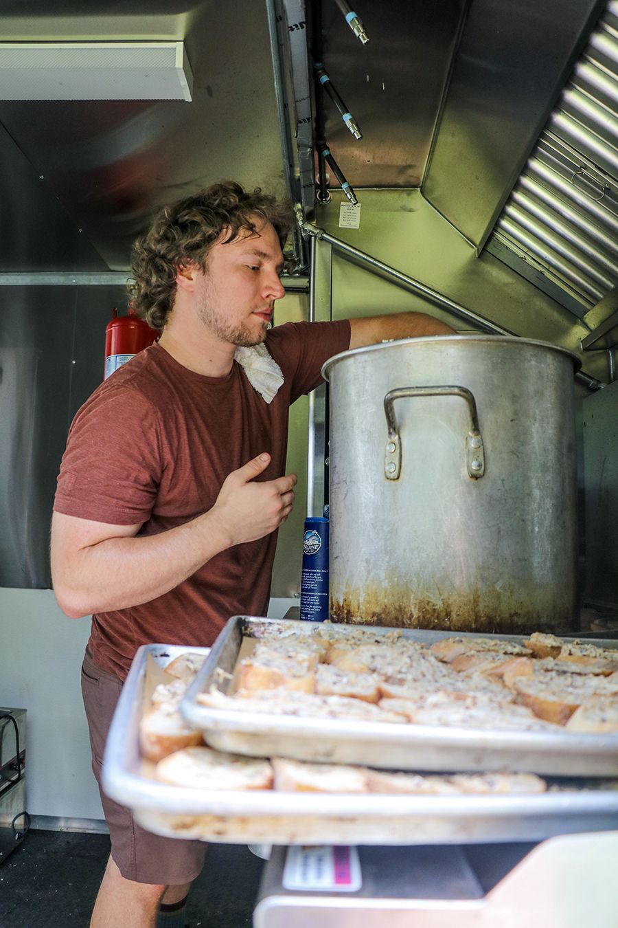 A chef stirs a large pot of grits in a kitchen with trays of appetizers stacked in the foreground.