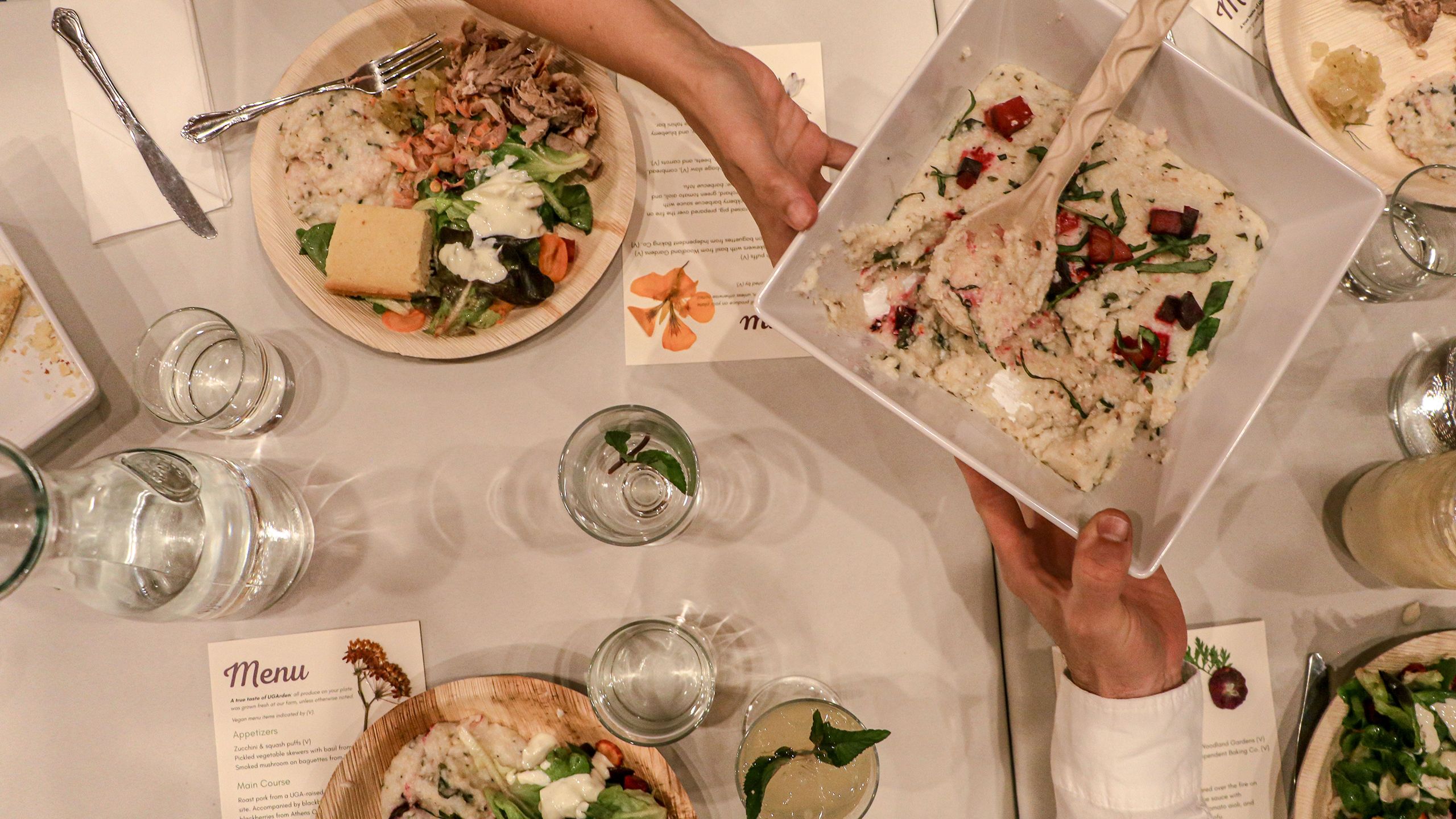 An overview photo of the place settings as dinner plates fill up with food and guests pass a white bowl of grits with red beets on top. 