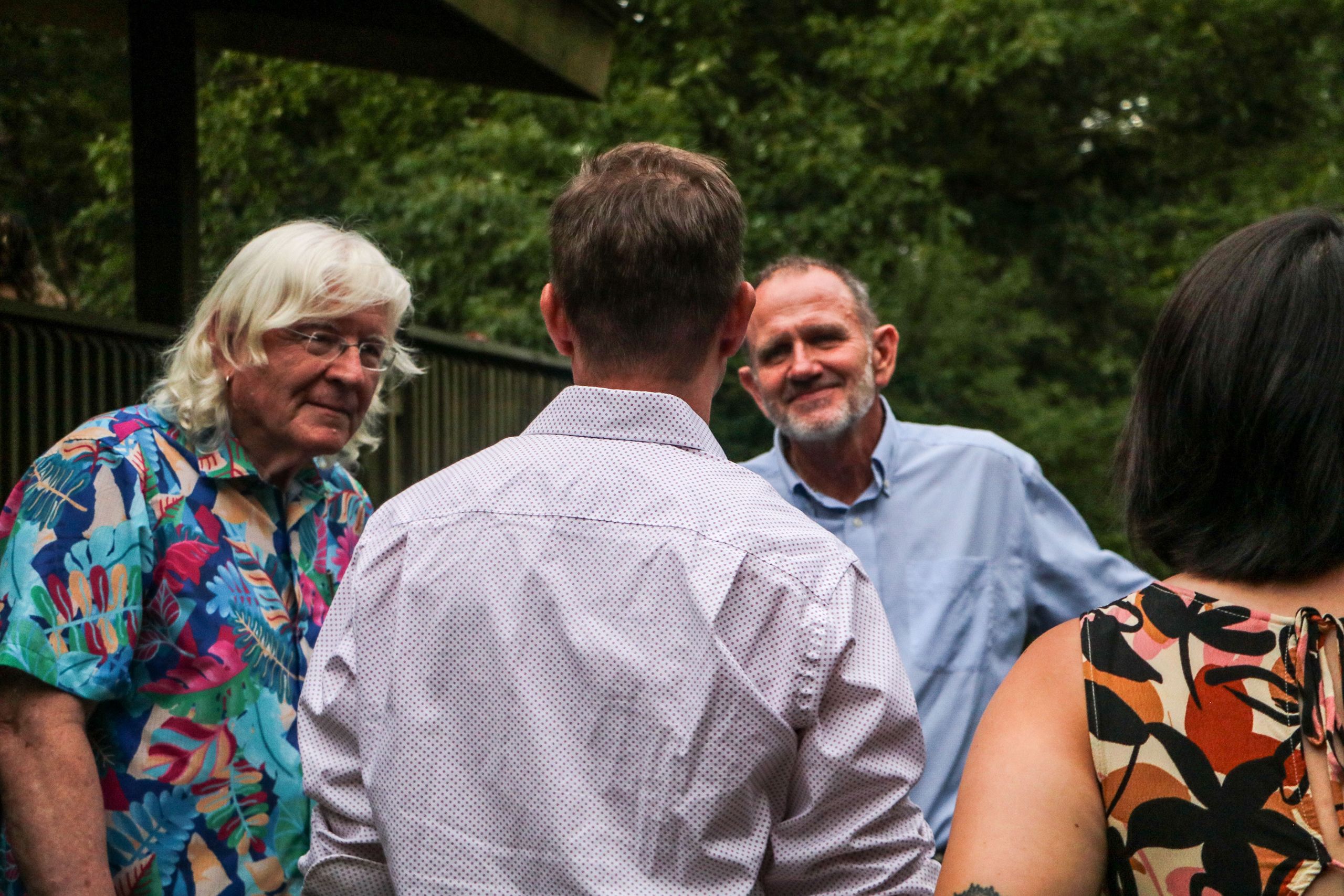 Three people talk amongst themselves. The man on the left has white hair and glasses and wears a colorful top. The man on the right has a blue shirt and is smiling. The man in the middle has his back facing the camera. 