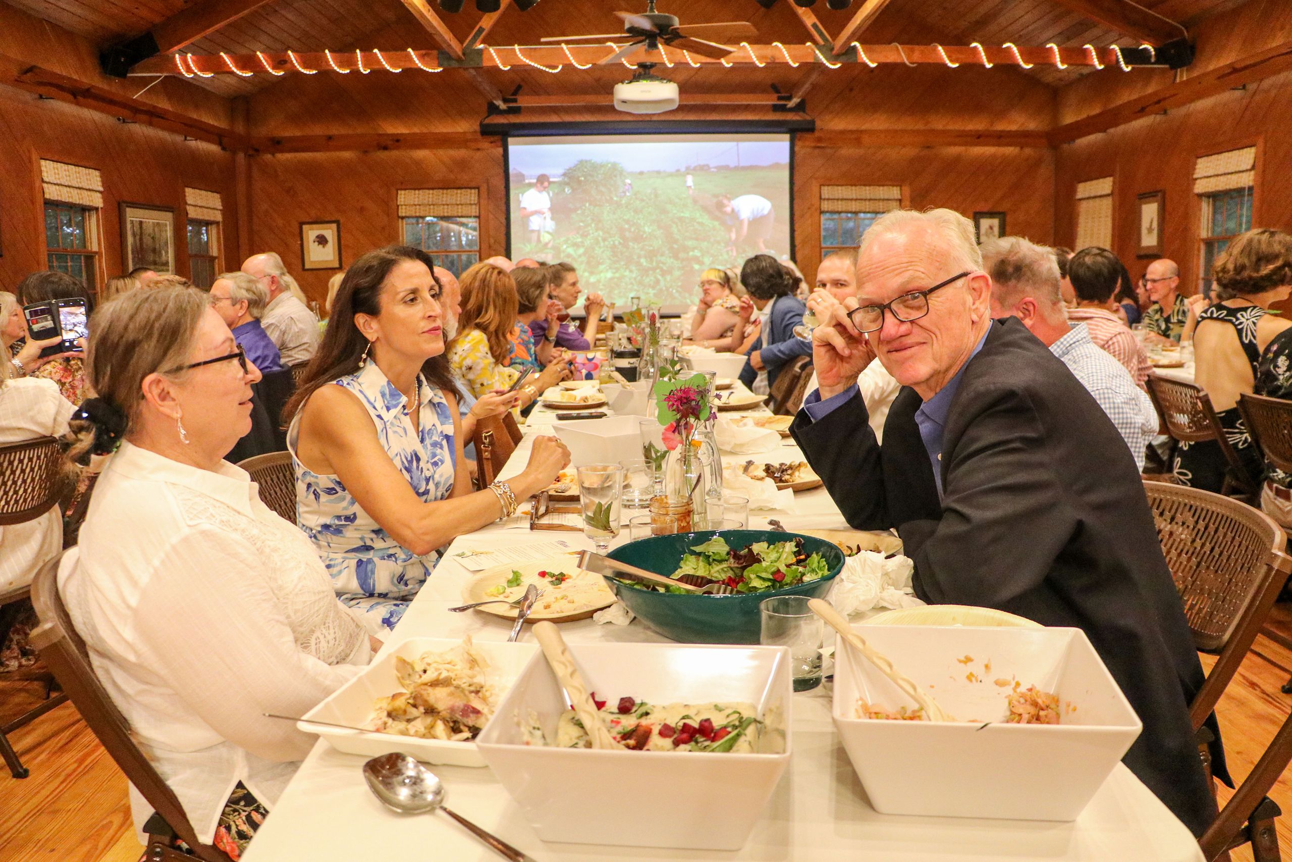 Looking down a long dinner table, guests are talking and the man in the front right wears glasses and looks at the camera and smiles. 
