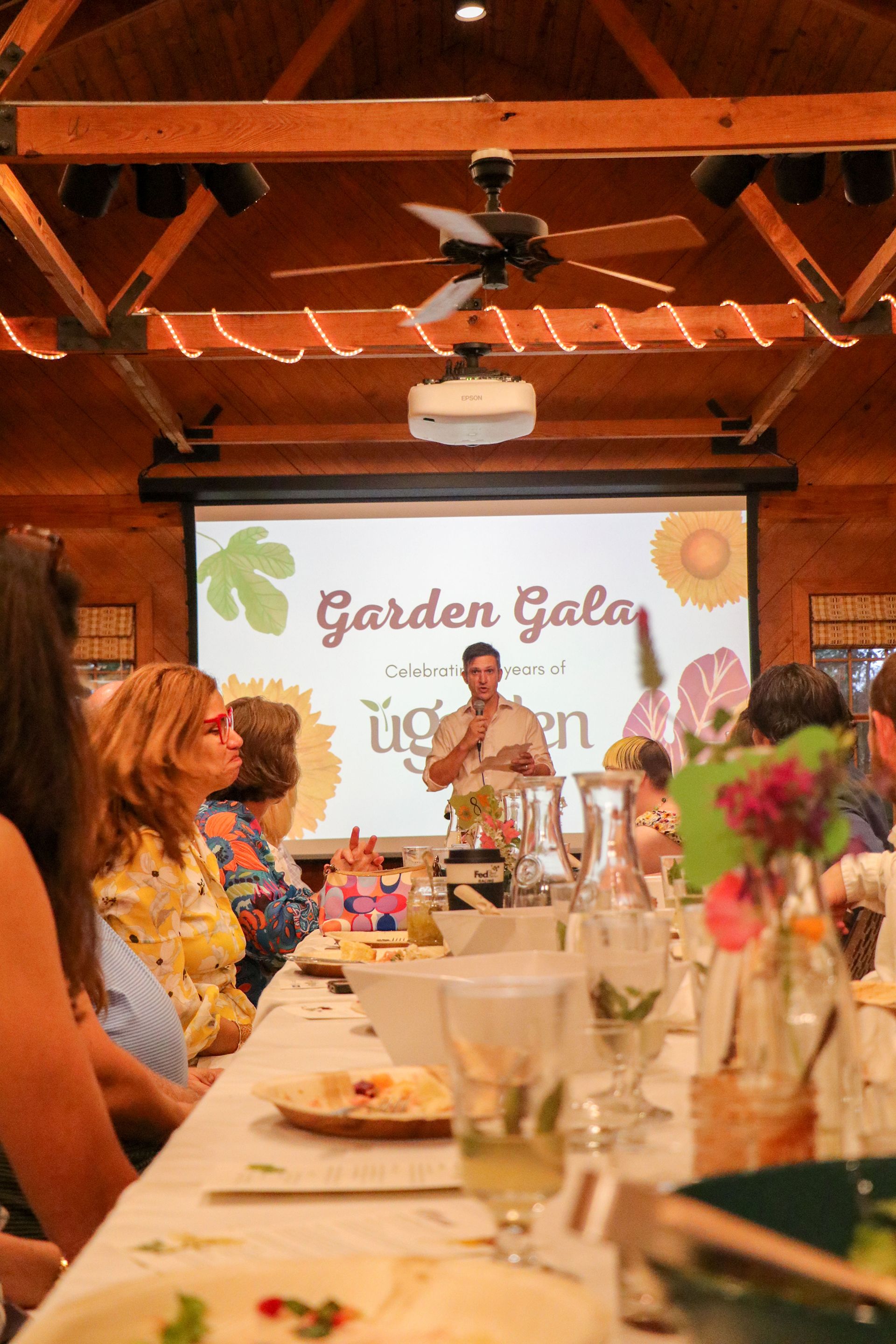 Ty Brooks holds the microphone and speaks to guests in front of a projector screen that says "Garden Gala"