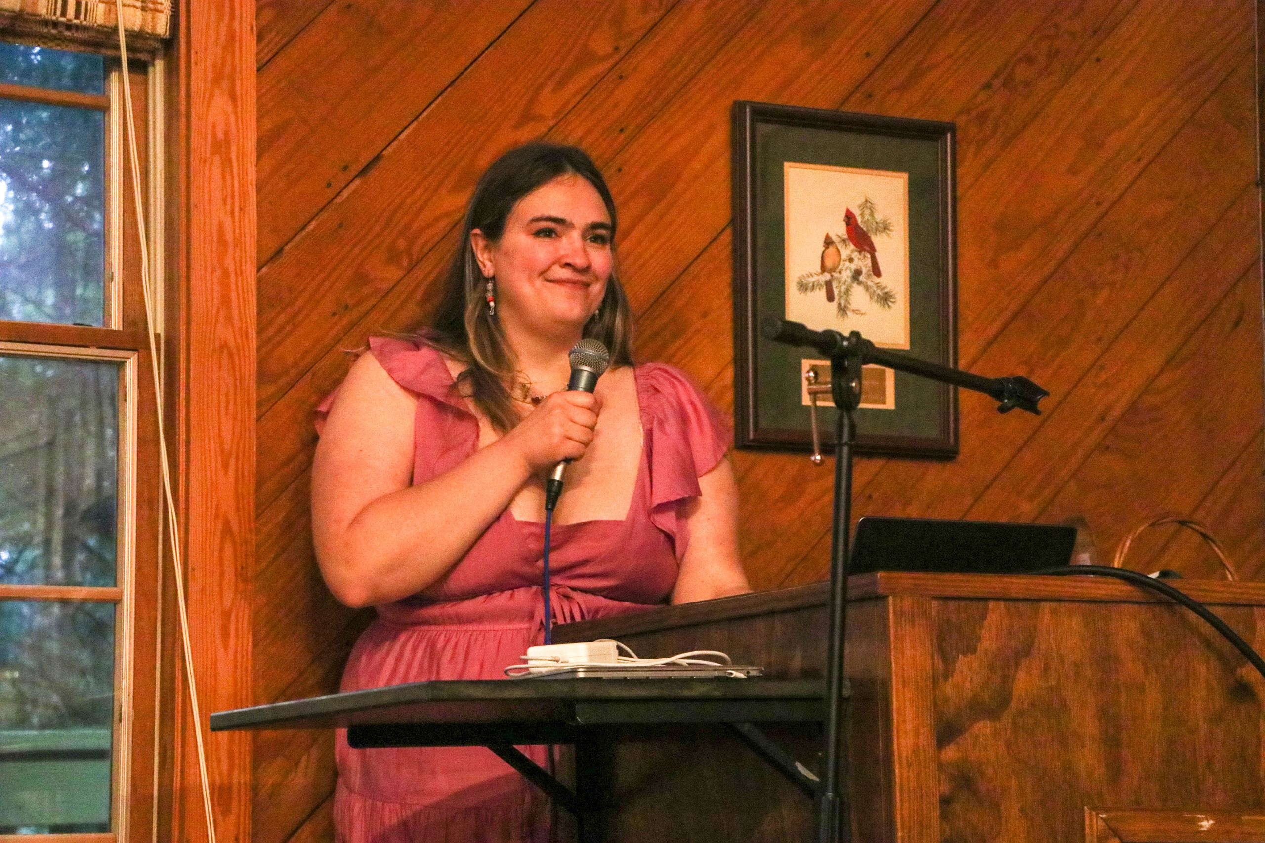 A woman in a pink dress stands at the lectern and holds a microphone as she talks to guests. 