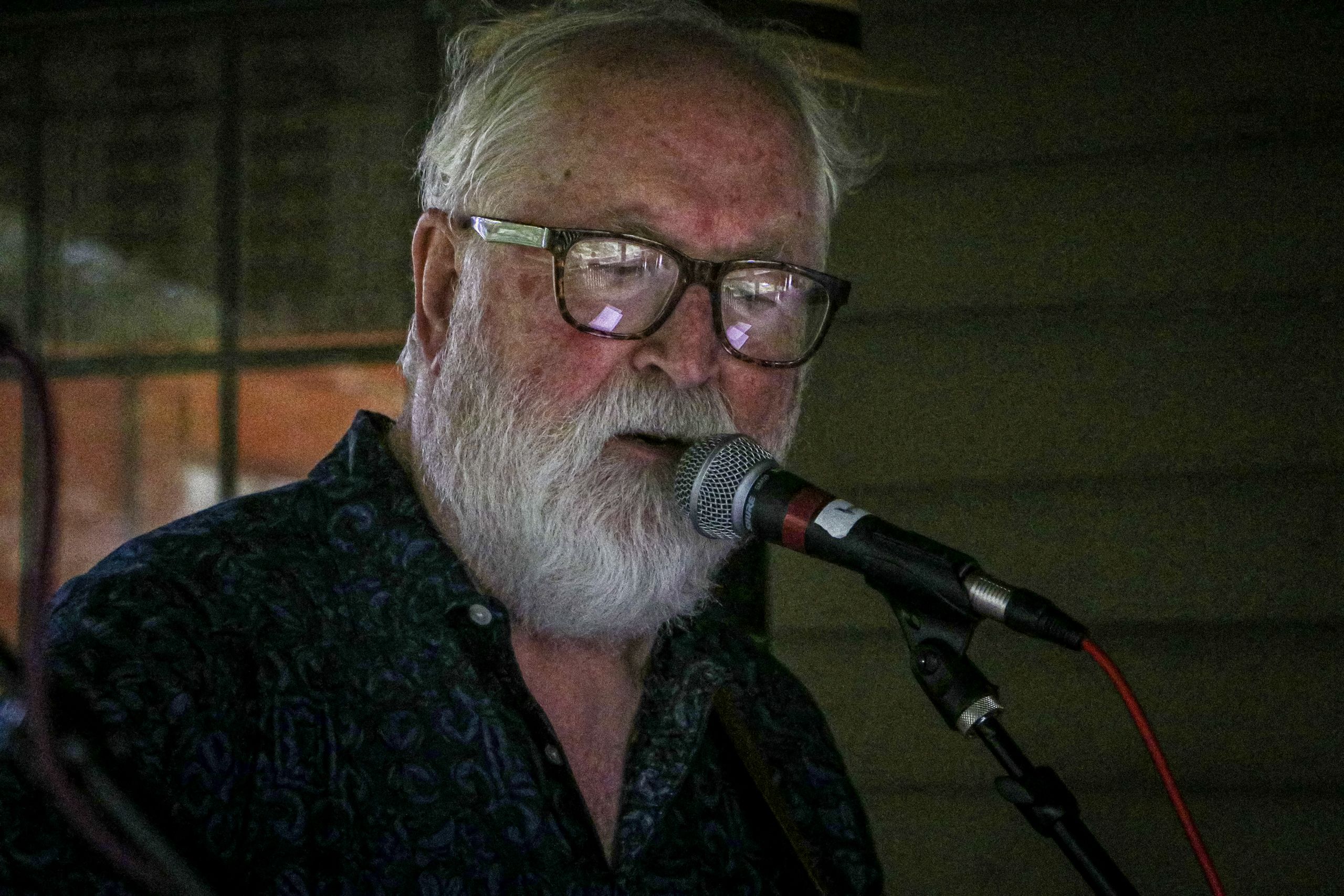 A close up image of a man with a white beard and glasses sings into the microphone on the covered deck. 