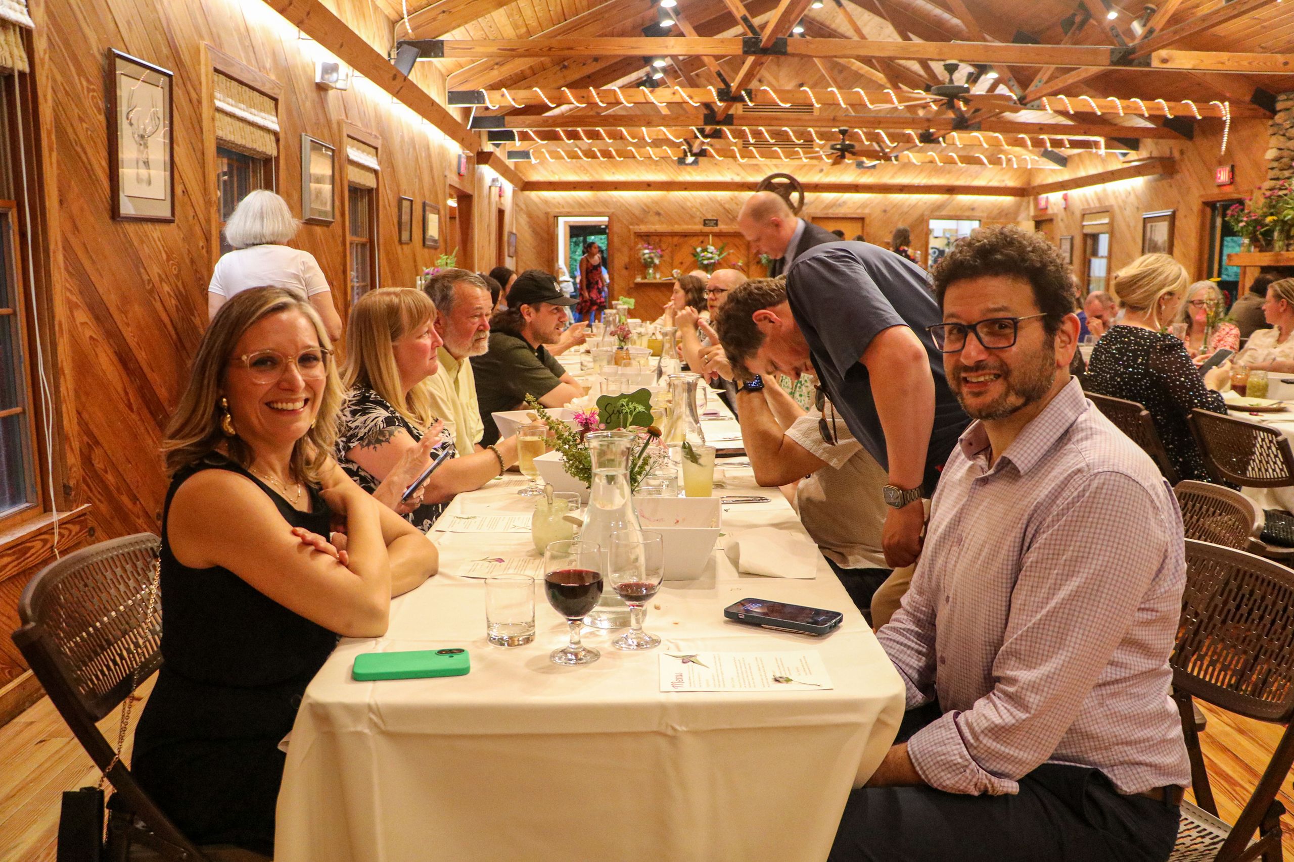 Looking down a long dinner table, guests are talking amongst themselves before dinner is served. The people in the front, both wearing glasses, smile at the camera. 