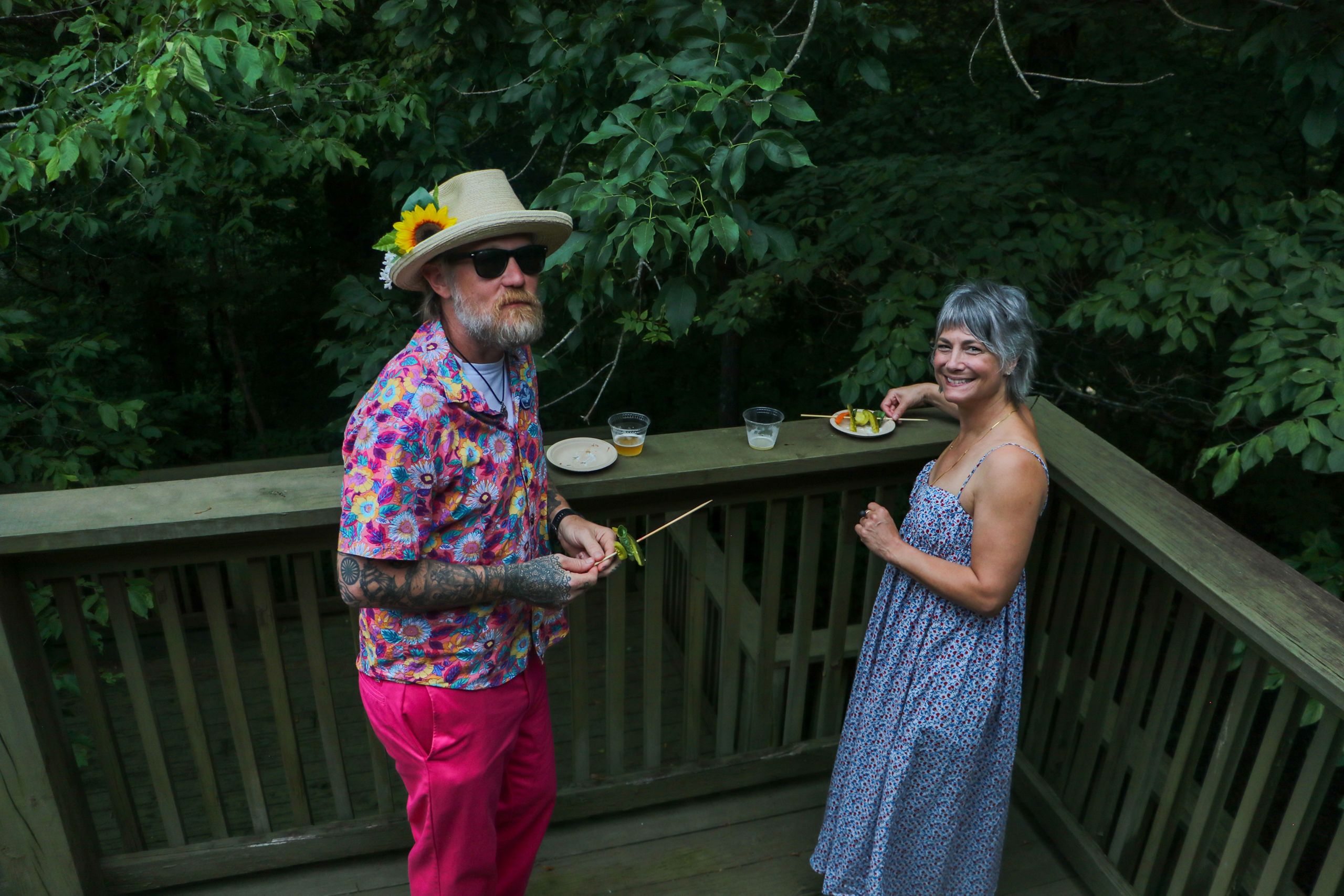 A man in a hat with sunglasses and a colorful, floral outfit stands on the deck with a woman on the right who wears a blue dress. Both are eating appetizers.