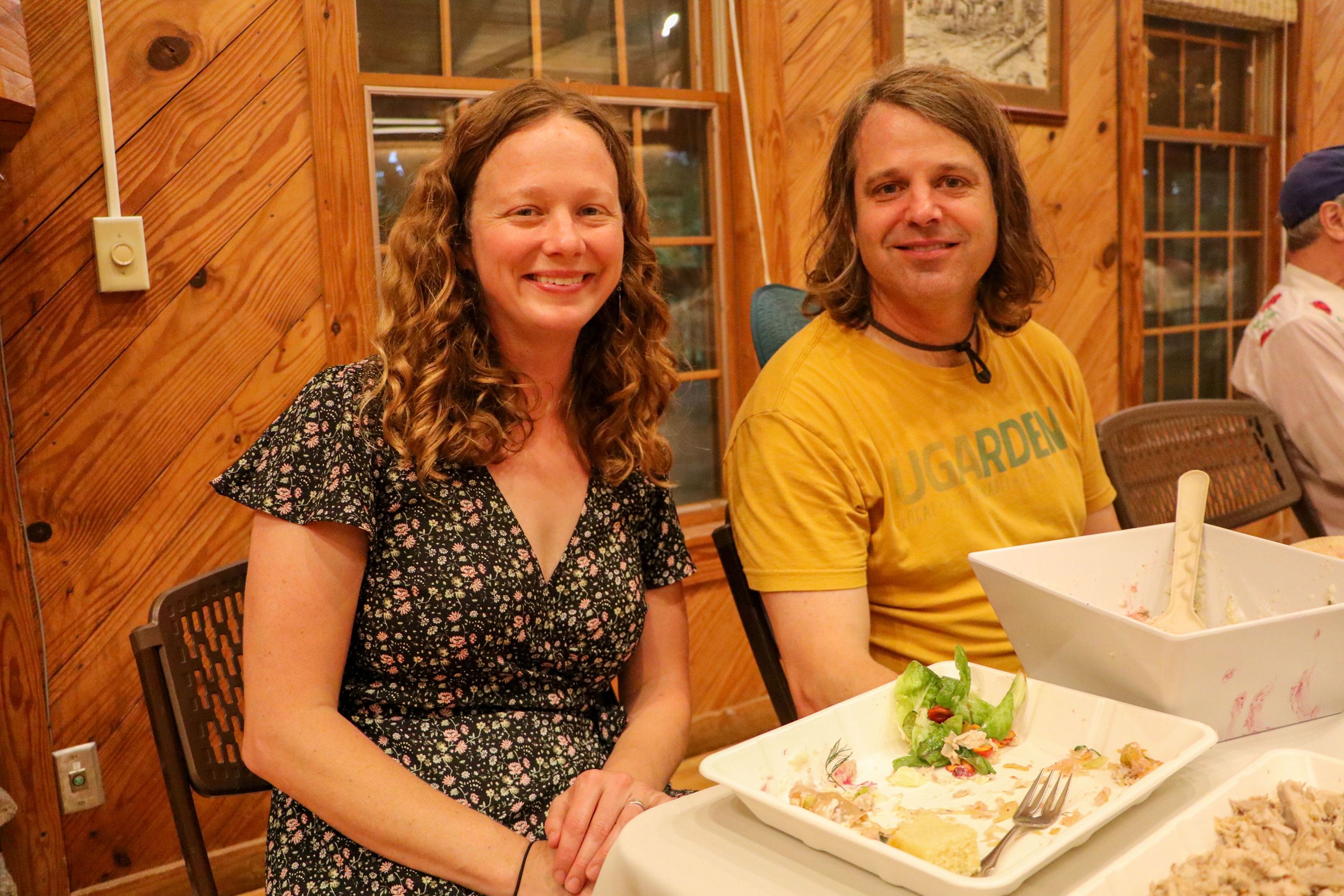 A woman wearing a blue floral dress smiles as she sits at the dinner table next to a smiling man in a yellow shirt.