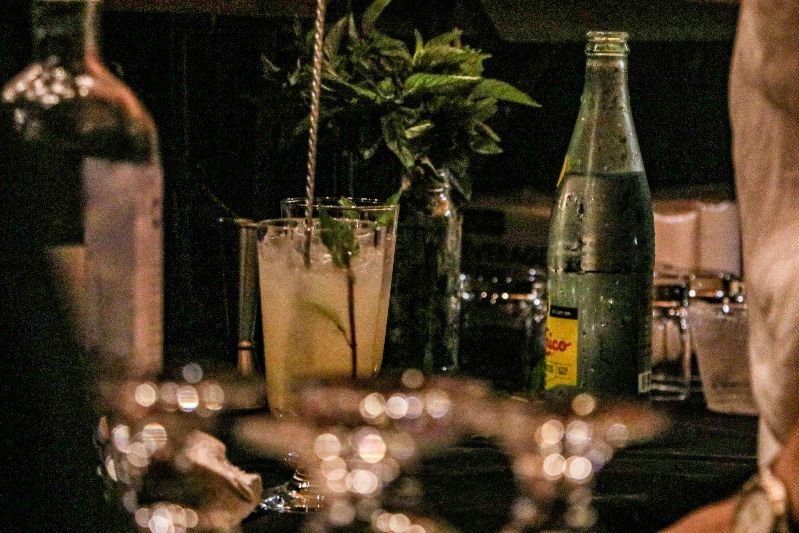 Specialty drinks are lined up at the bar with herbs and seltzer water nearby.