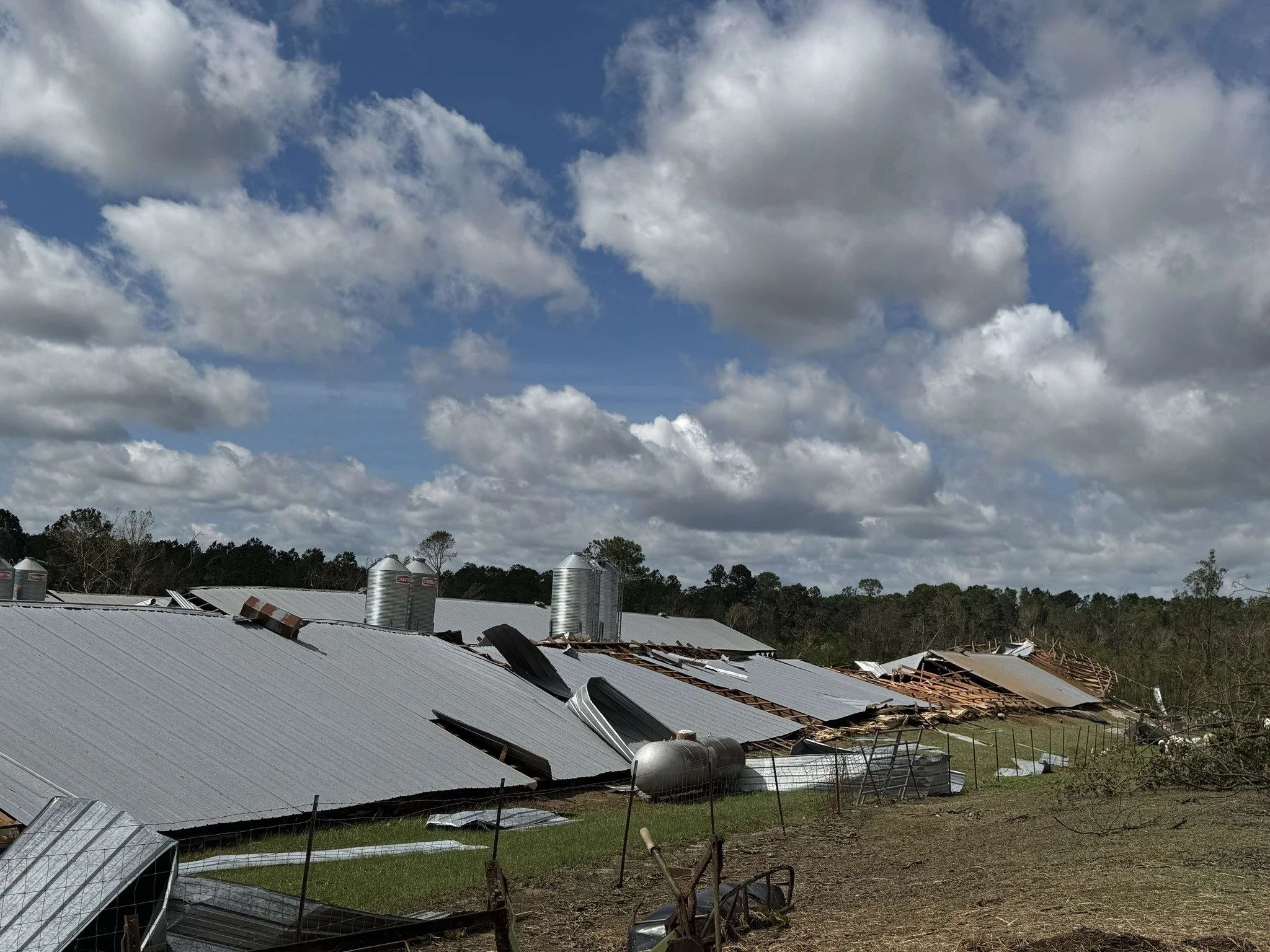 Damaged farm buildings with torn-off metal roofing and exposed wooden frames, set against a partly cloudy sky.