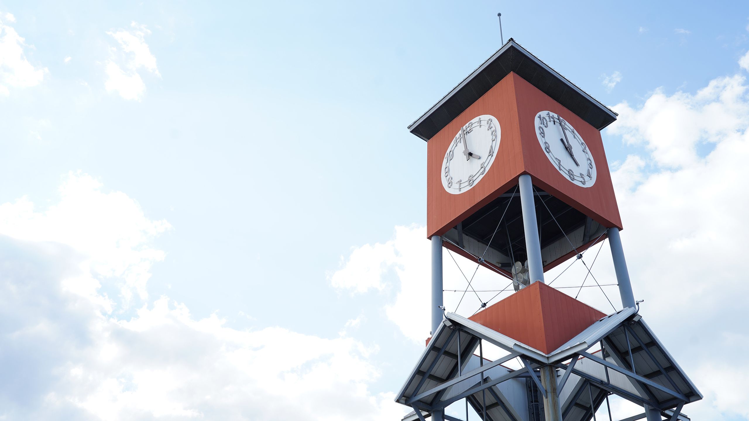 Red clock tower with white clock faces showing time, supported by metal beams, against a blue sky with scattered clouds.