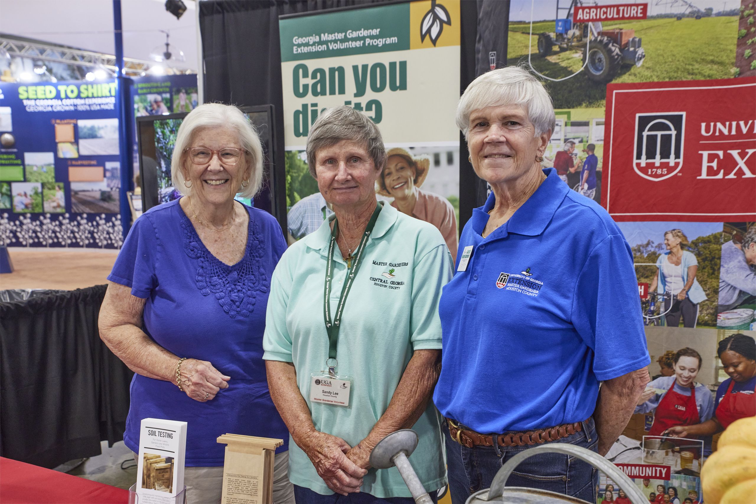 Three women stand together at a gardening exhibit, wearing volunteer shirts, with agriculture and extension program banners in the background.