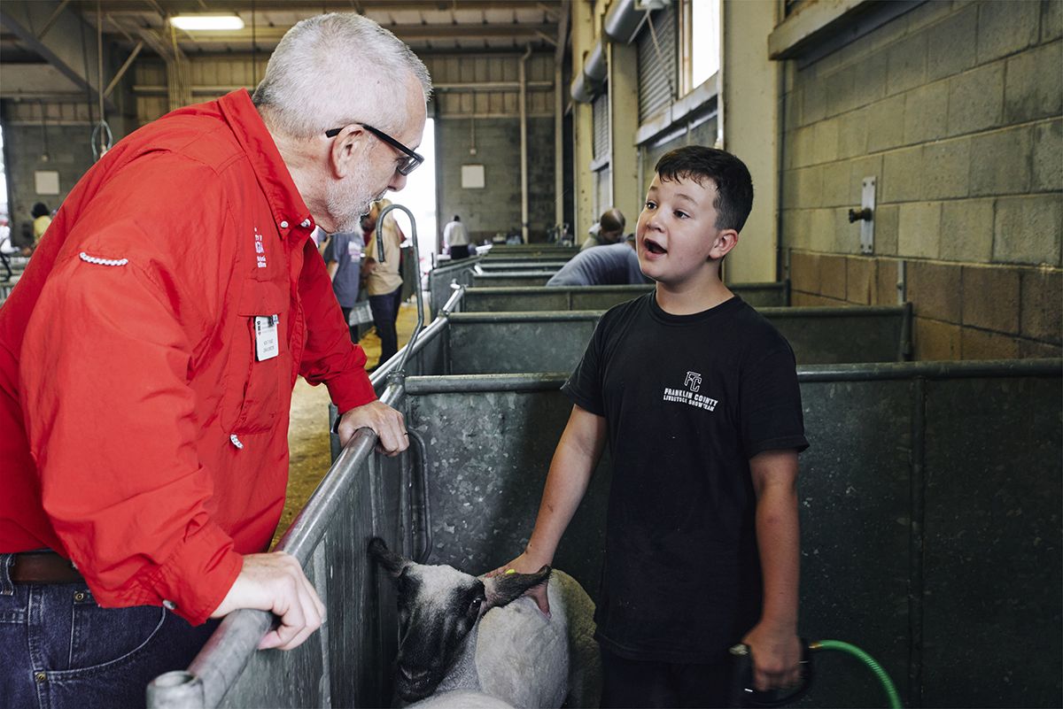 Man in red jacket and boy in black shirt interact with a sheep in a barn, leaning over a metal pen.