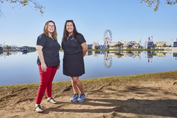 Two women stand by a lake with a fairground and Ferris wheel reflected in the water behind them.