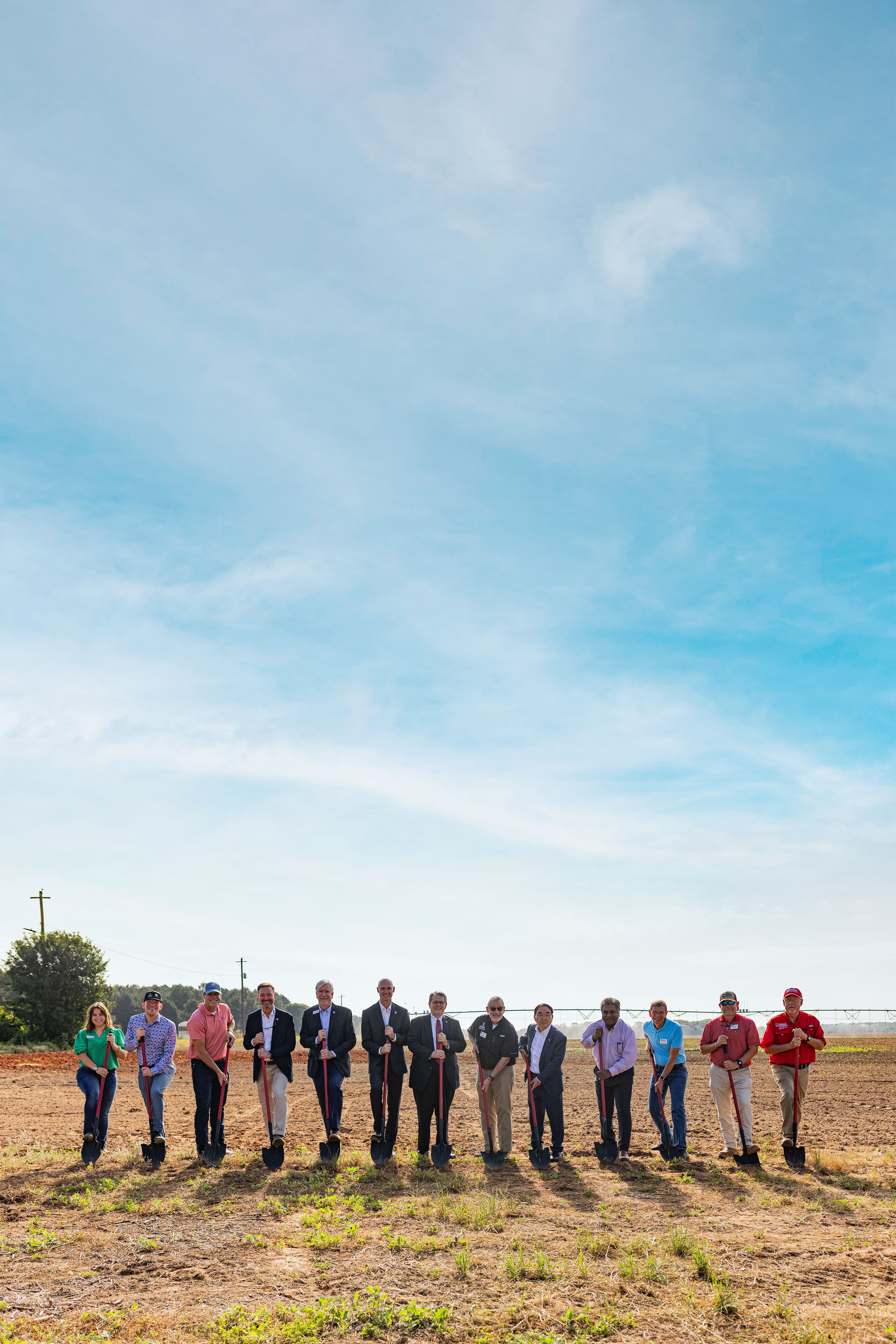 A diverse group of people stands in a field under a clear blue sky, each holding a shovel for a groundbreaking ceremony, conveying a sense of community and progress.