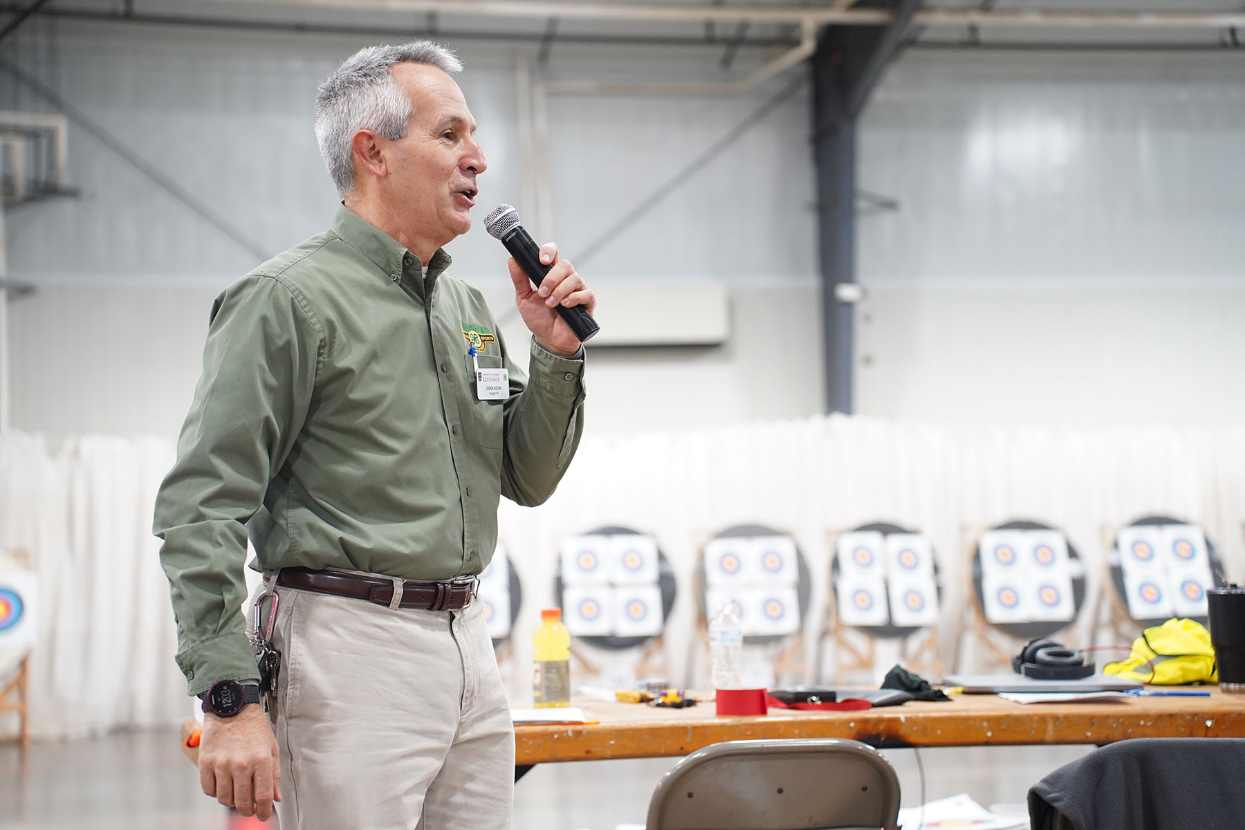 A man in a green button down shirt speaks into a microphone with a tabel and archery targets in the background.