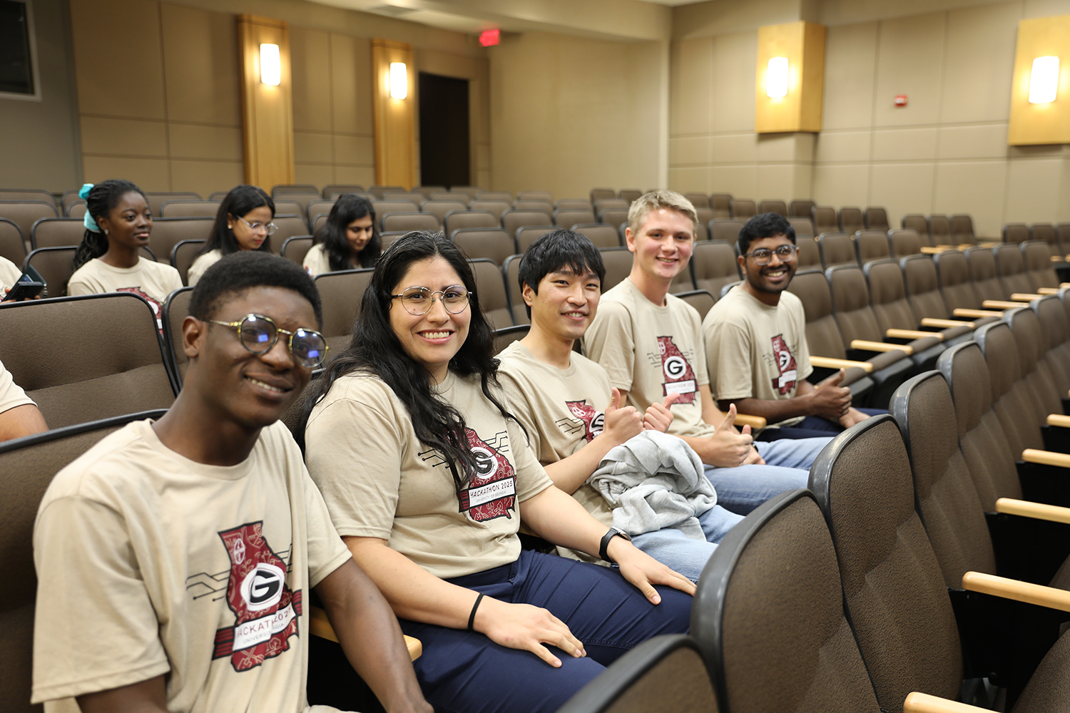 five students smile for the camera while sitting in a row of brown theater seats.